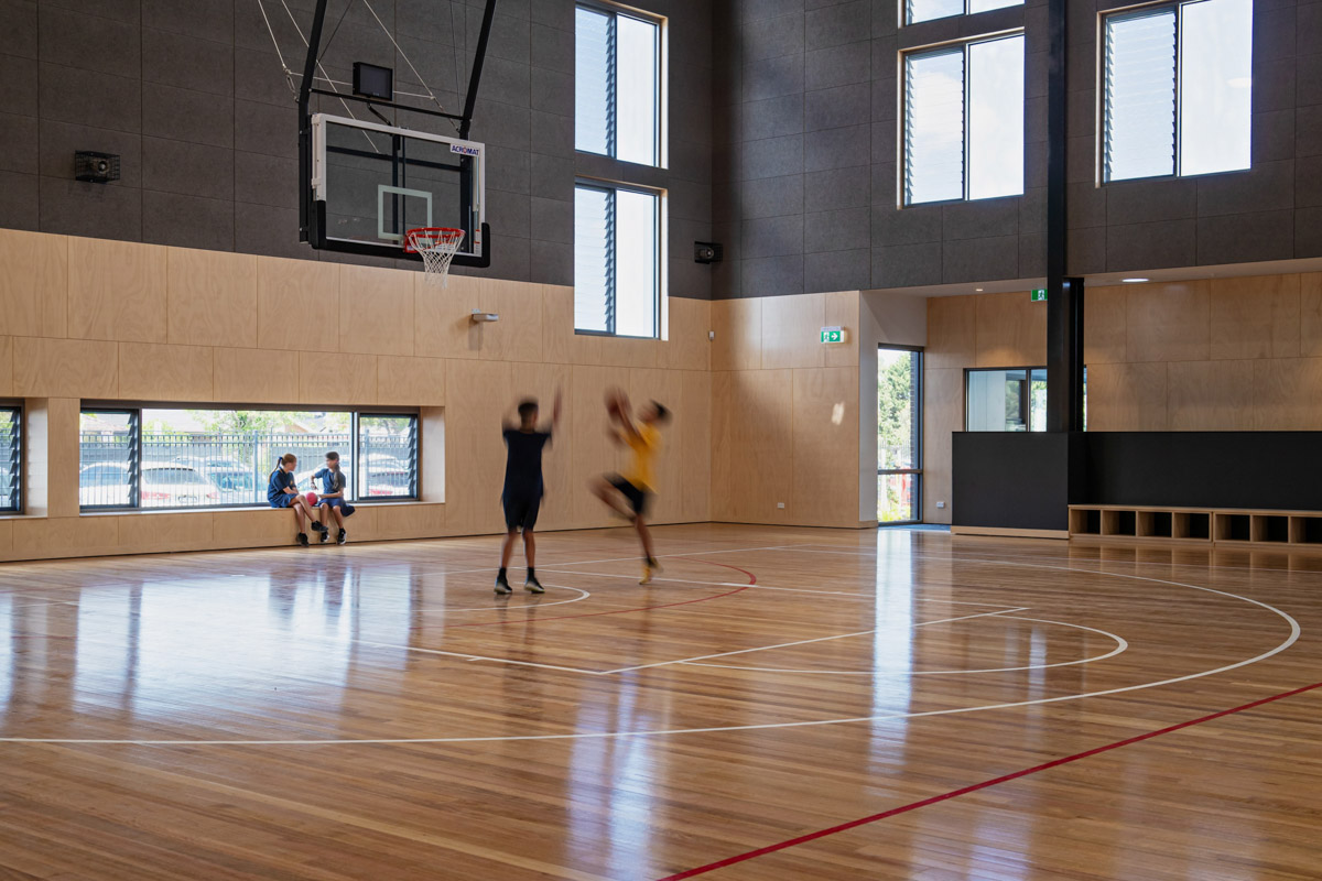 Two students playing basketball on a polished timber indoor court with a hoop mounted above. Large windows and timber wall panels surround the space.
