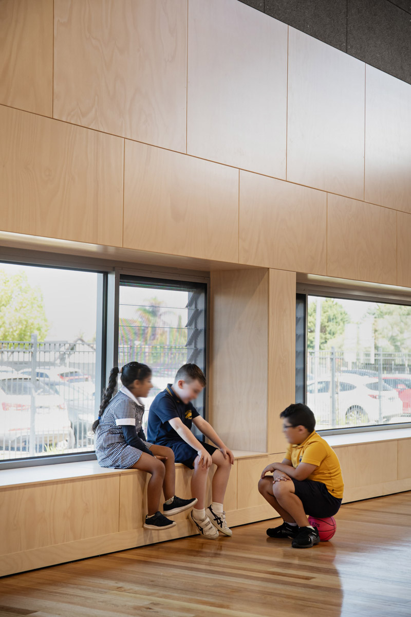 Three students sitting on a timber bench built into a wall with large windows overlooking the outdoor area. The floor is polished timber.