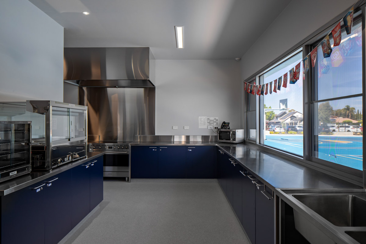 A school kitchen with stainless steel benches, deep sinks, and blue cabinetry. Large windows overlook the outdoor sports courts, and commercial appliances are installed along the walls.