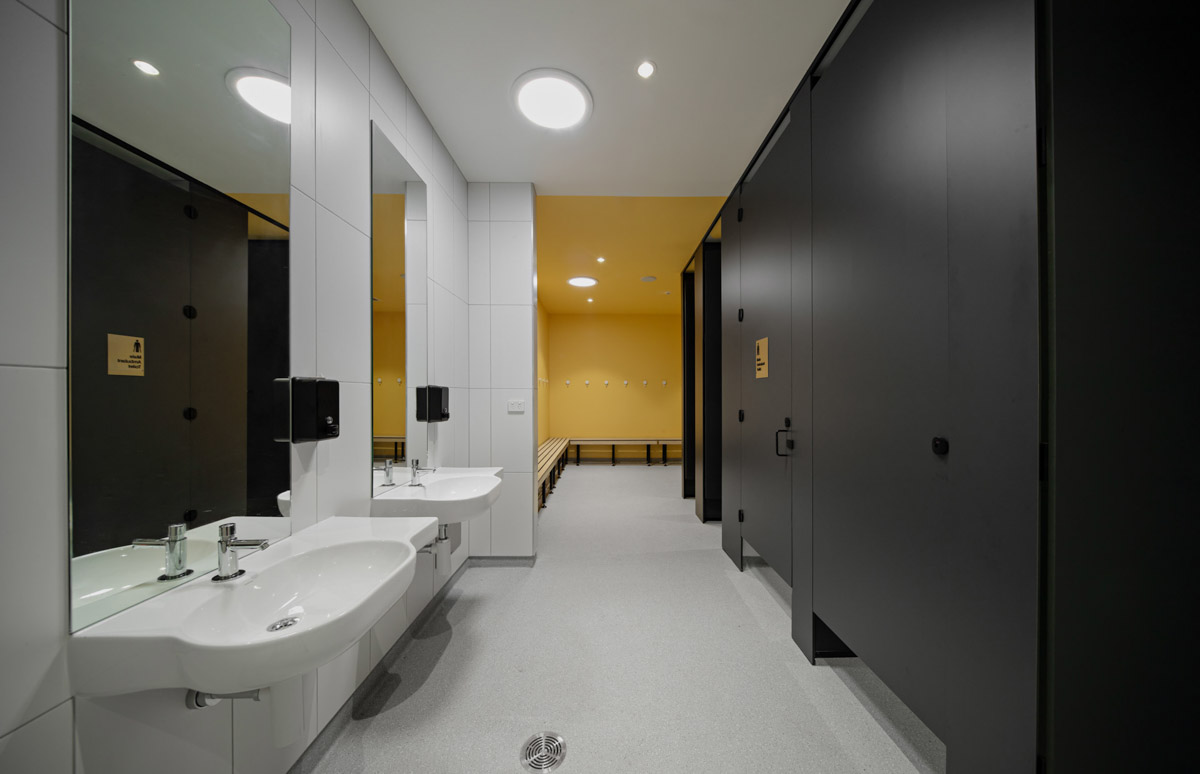 A modern student bathroom with white tiled walls, two basins with mirrors, black cubicle doors, and a yellow-painted change area with timber benches and hooks.