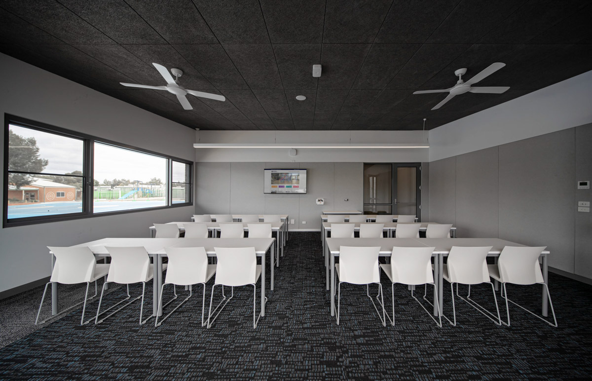 A classroom with rows of white chairs and tables facing a wall-mounted screen. Large windows provide natural light, and the room has dark acoustic ceiling panels and fans.