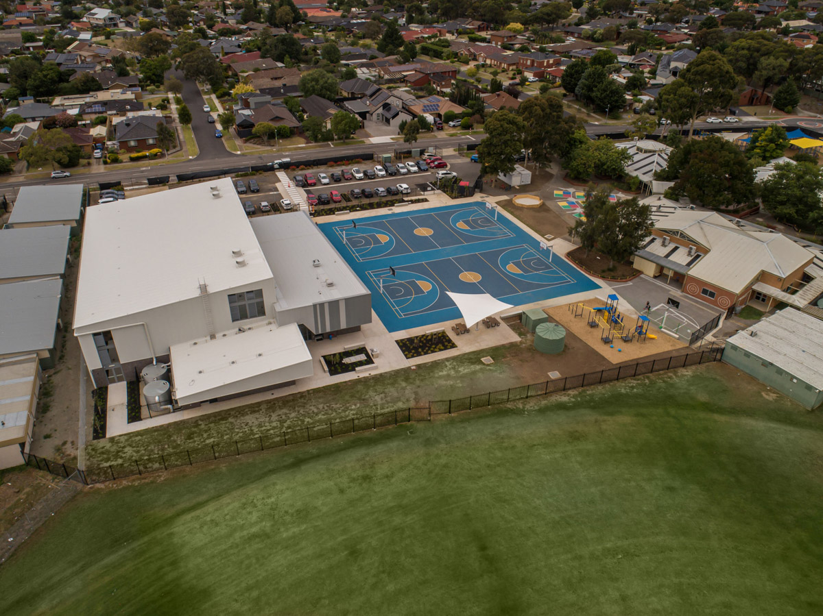 An aerial view of a school campus showing a large building with a white roof, multiple blue outdoor sports courts, landscaped areas, and surrounding residential streets.