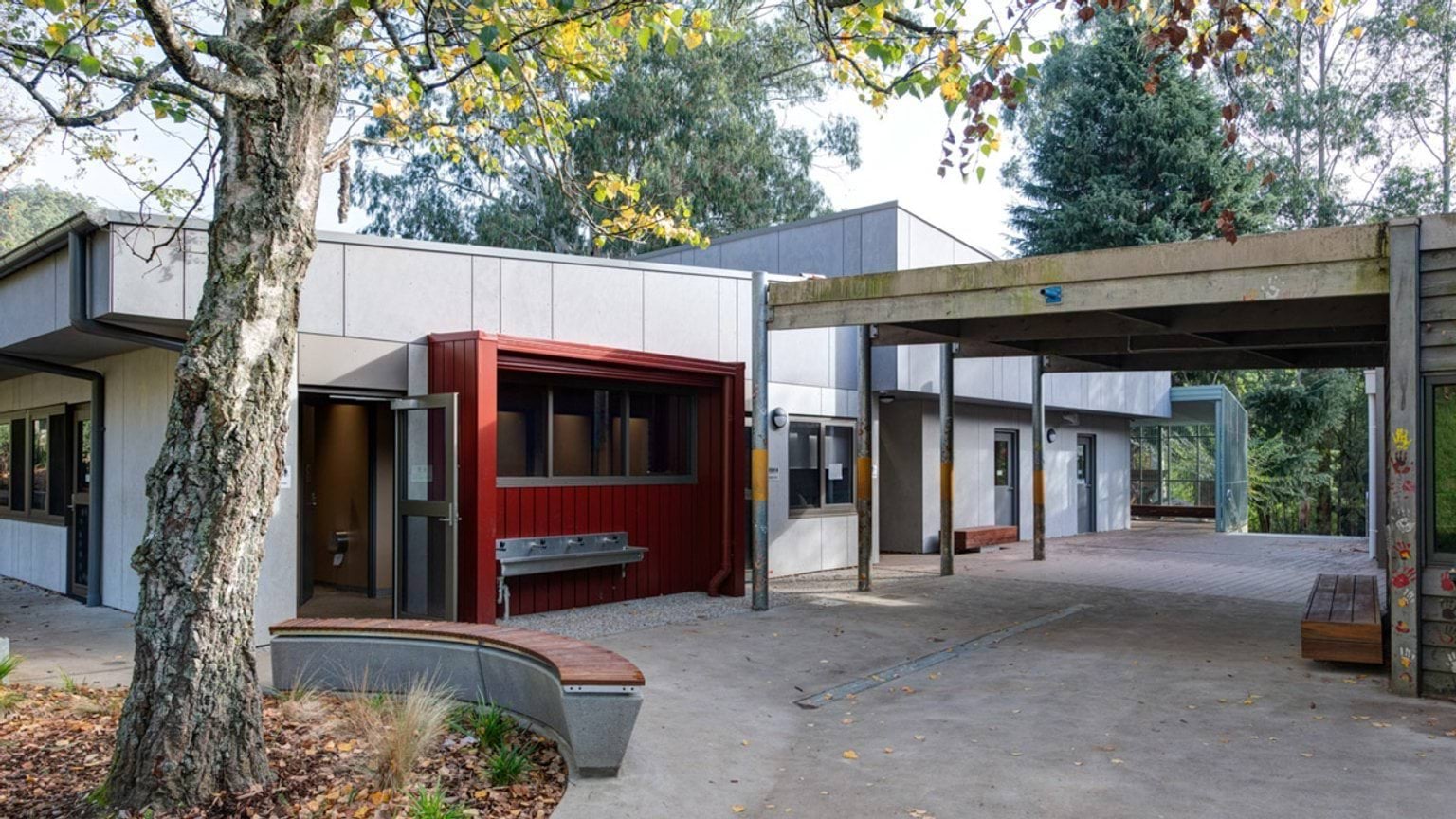 A school courtyard with grey cladding, a red feature wall, and a stainless steel drinking trough. Timber benches and garden beds are arranged under a covered walkway.