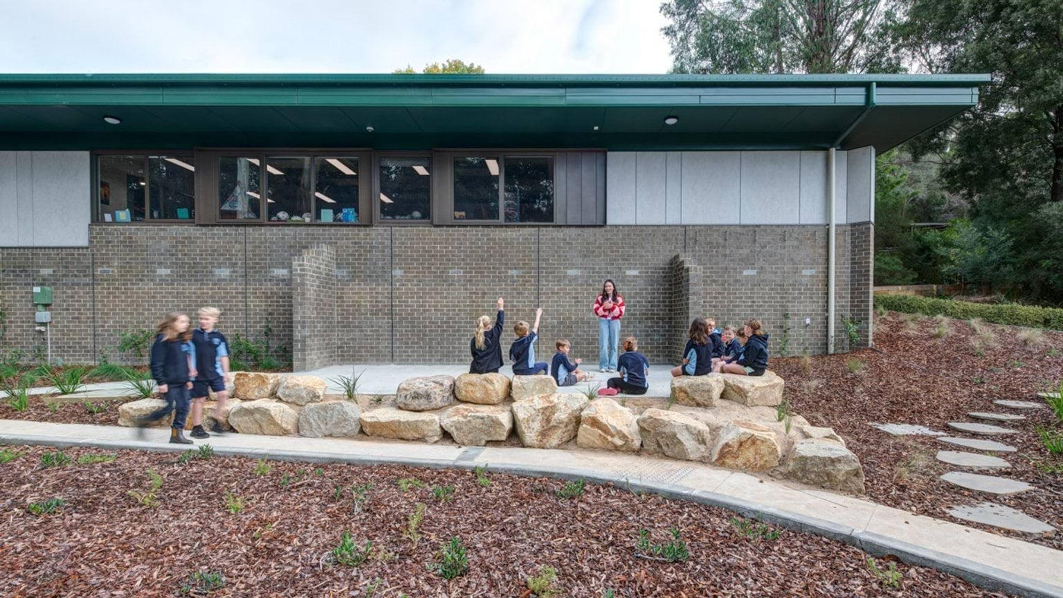 Students sitting on large rocks in a landscaped outdoor learning area with garden beds. A teacher stands nearby, and the school building with green roof is in the background.