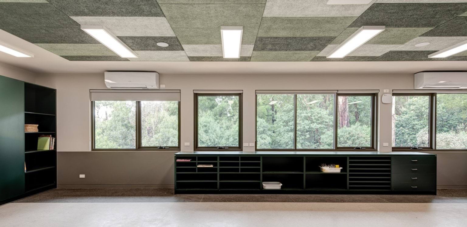 A classroom with large windows overlooking trees, green cabinetry with open shelving, and patterned green ceiling panels with LED lights.