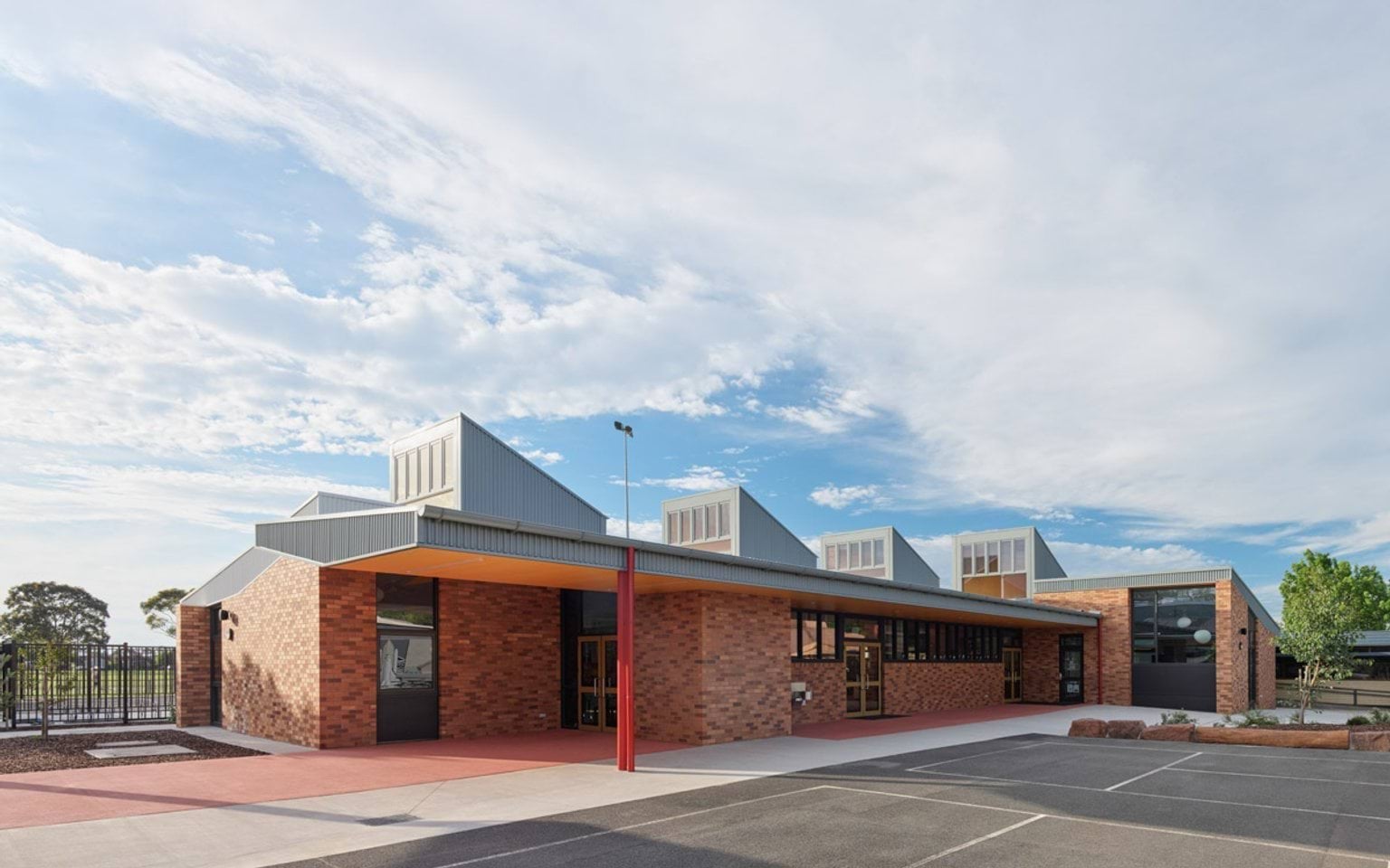 Modern brick school building with angled metal roof sections and large windows under a bright blue sky.