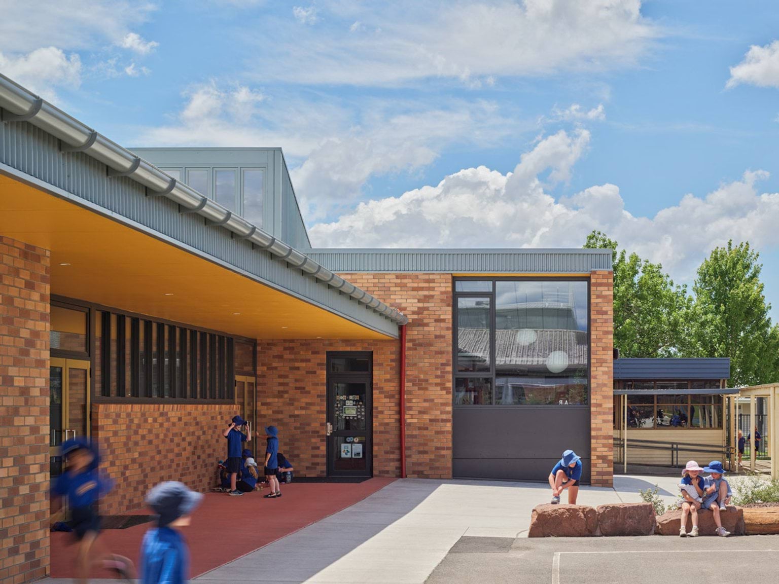 Students sitting and playing near a brick school building with yellow canopy and large windows.