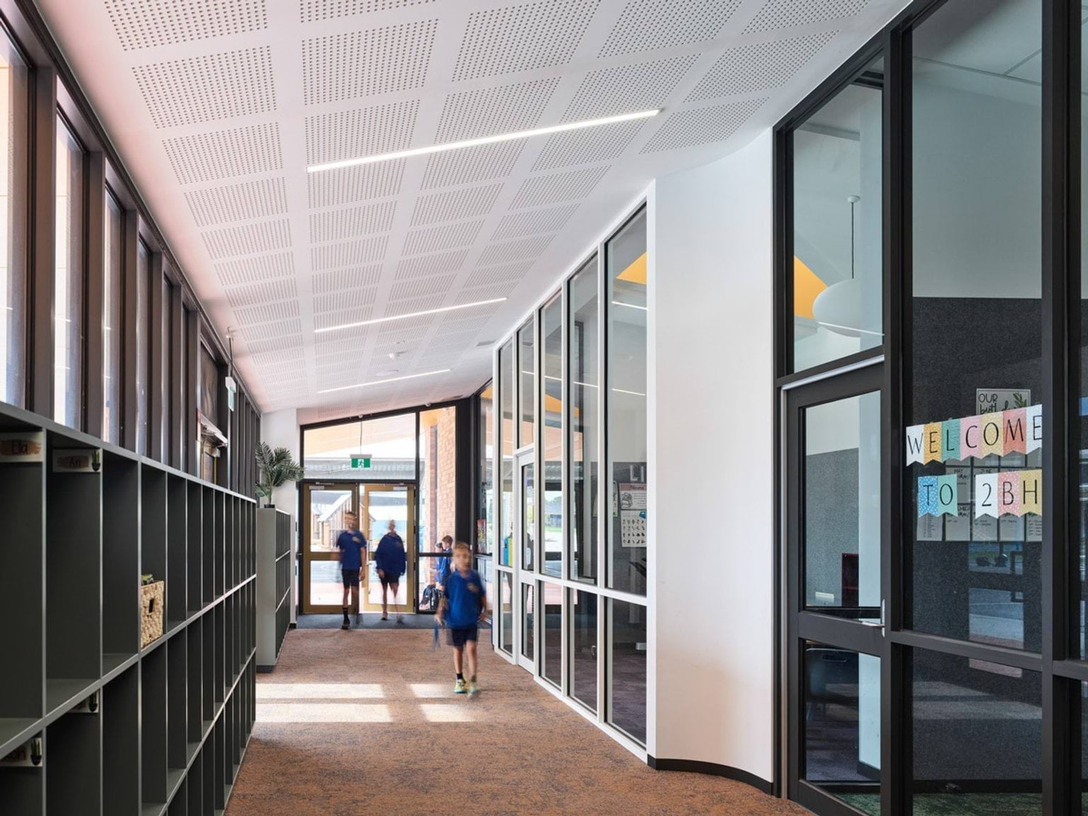 Bright school corridor with glass walls, cubby storage, and a colourful welcome sign on a classroom door.