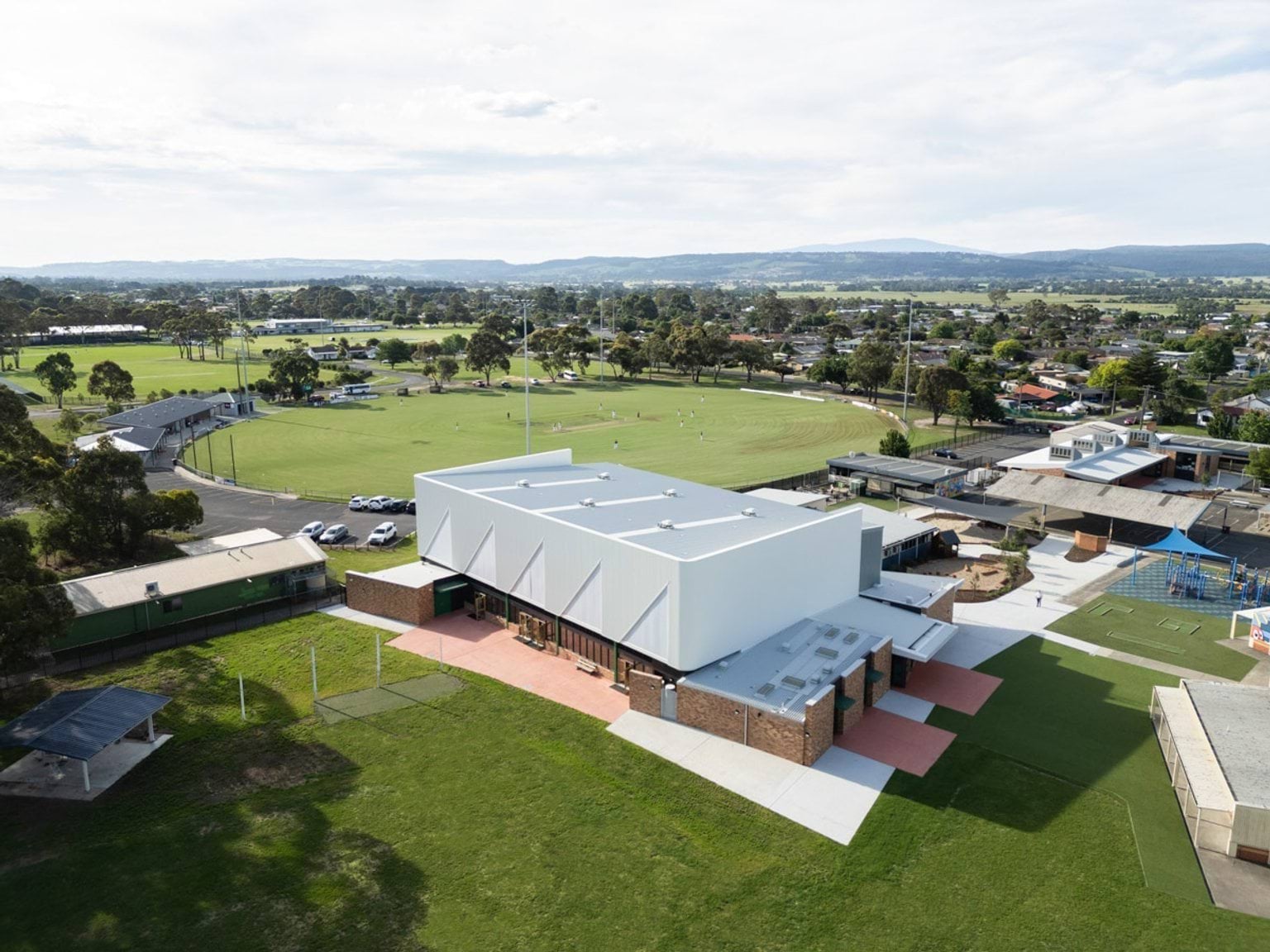 Elevated view of school sports hall and surrounding oval with suburban landscape in the background.