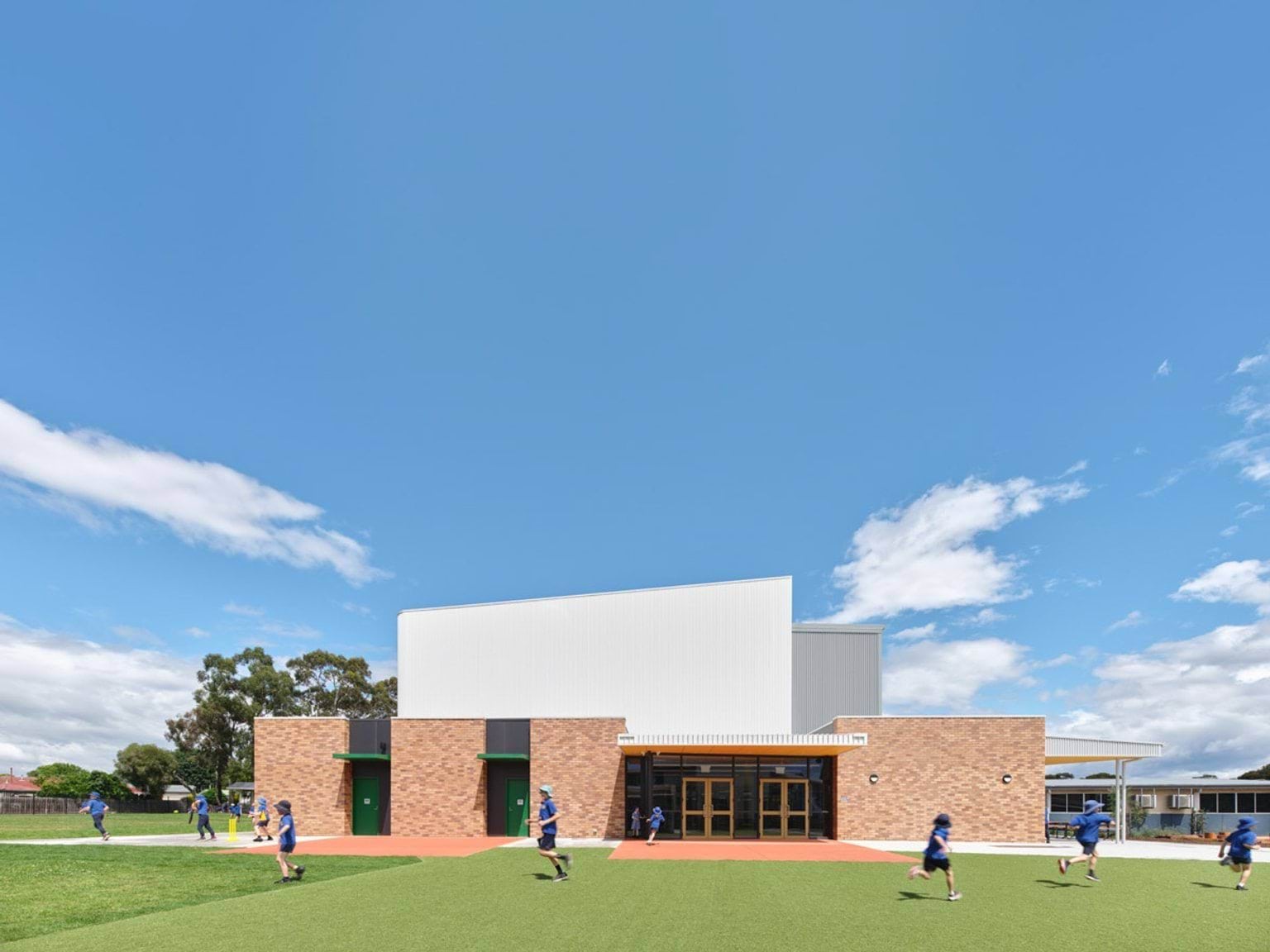 School sports hall with brick and white exterior, children running on synthetic turf under a clear sky.