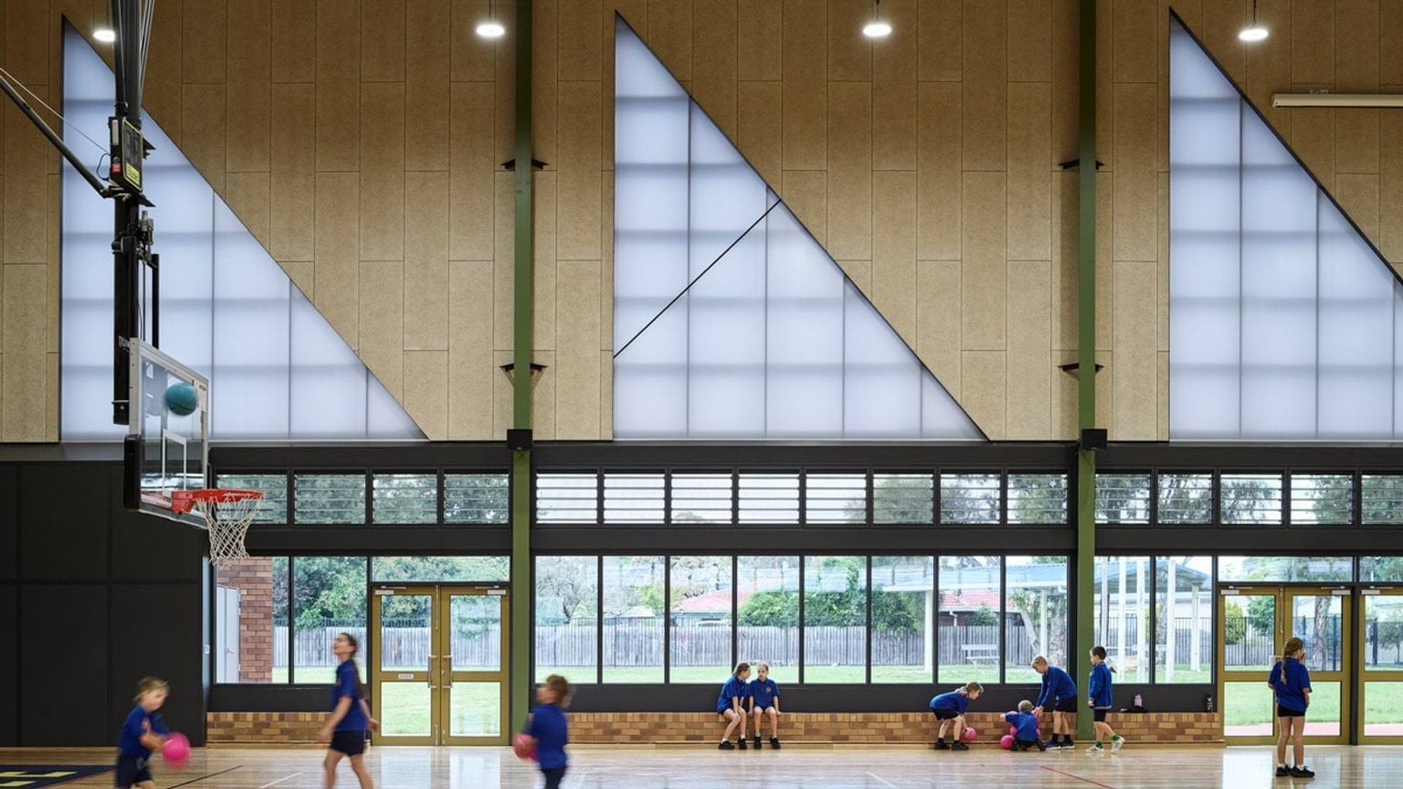 Interior of school gym with timber floor, triangular translucent panels, and students playing basketball.