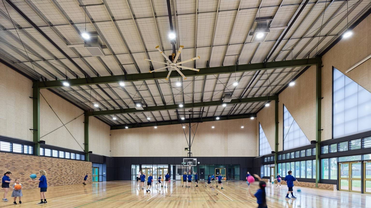 Spacious school gym with timber flooring, green steel beams, and students playing ball games.