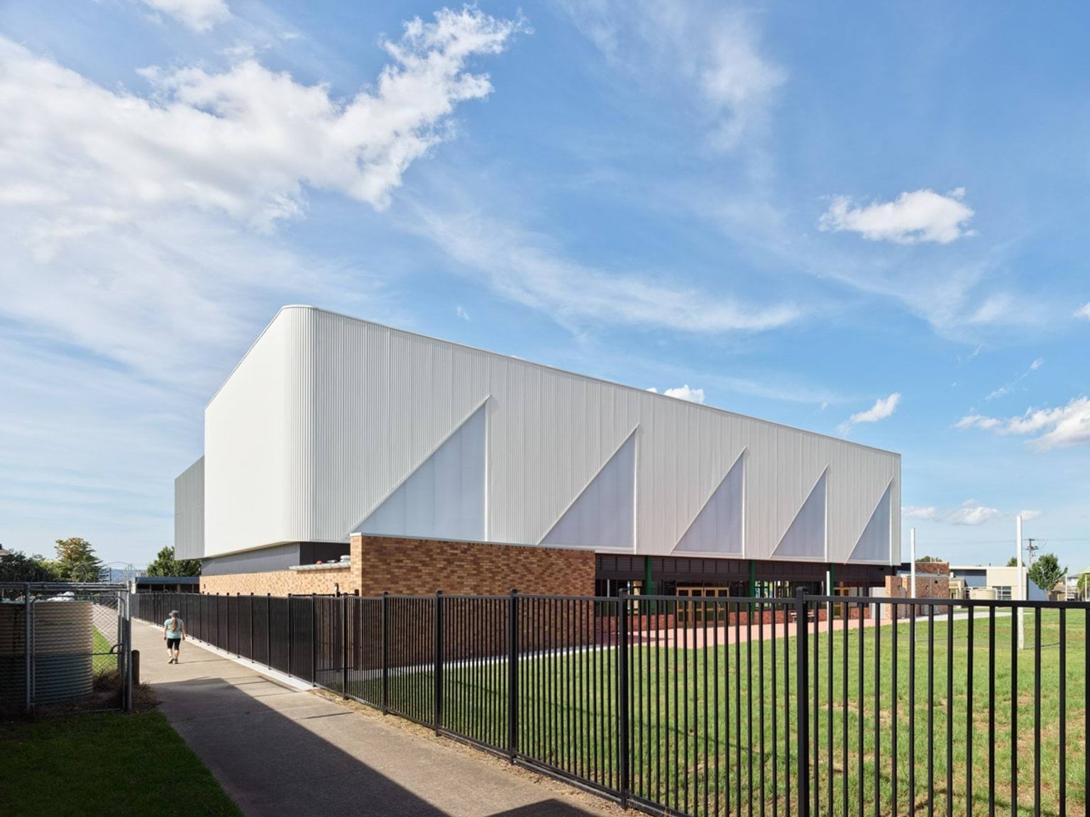White-clad school sports hall behind a black metal fence, with brick base and open grassy area.