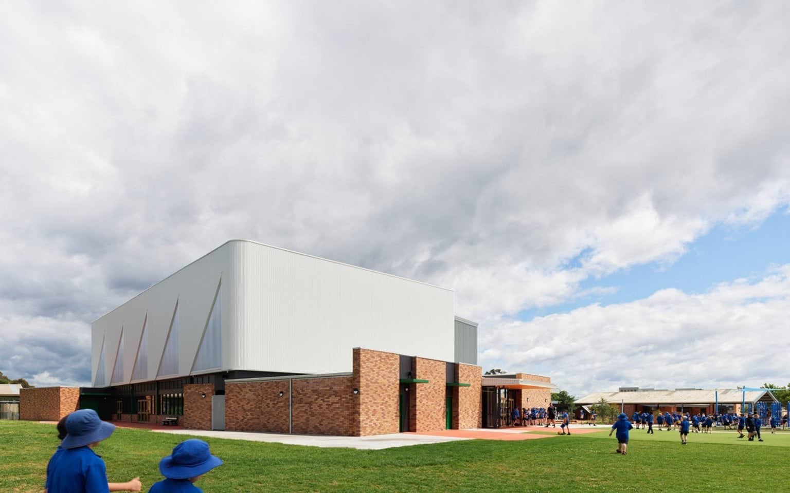 Large school sports hall with brick base and white cladding, students playing on a green field.