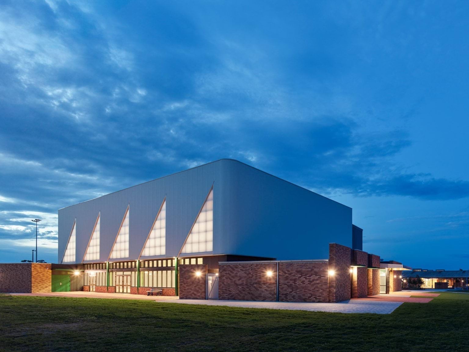 School sports hall illuminated at dusk, featuring brick walls and white cladding with triangular panels.