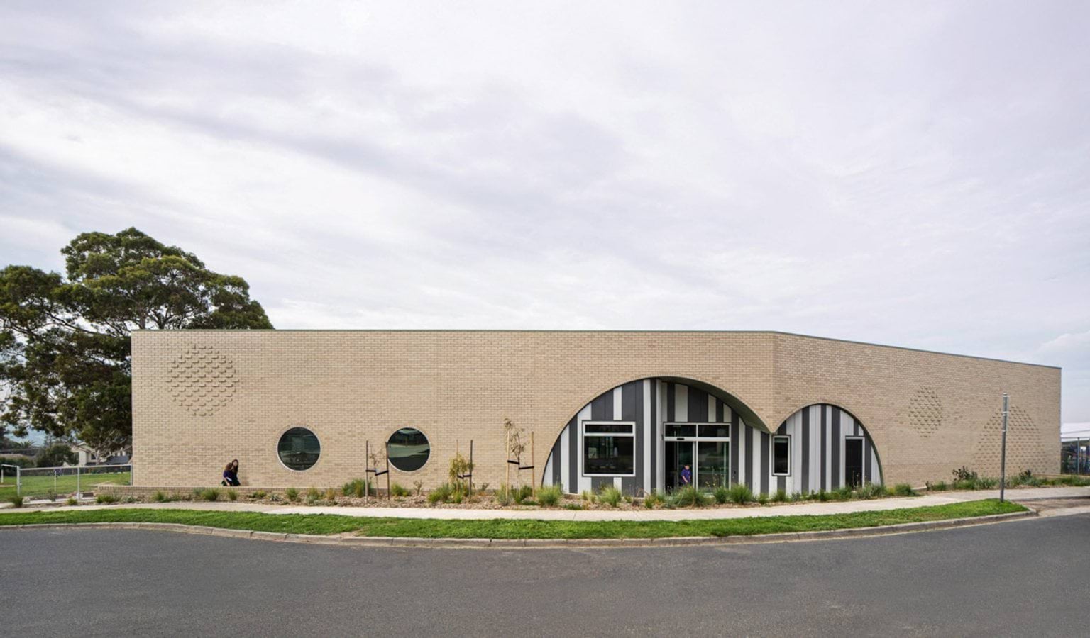 Exterior of a school building with light brick walls, circular windows, and arched entrances framed by striped panels.