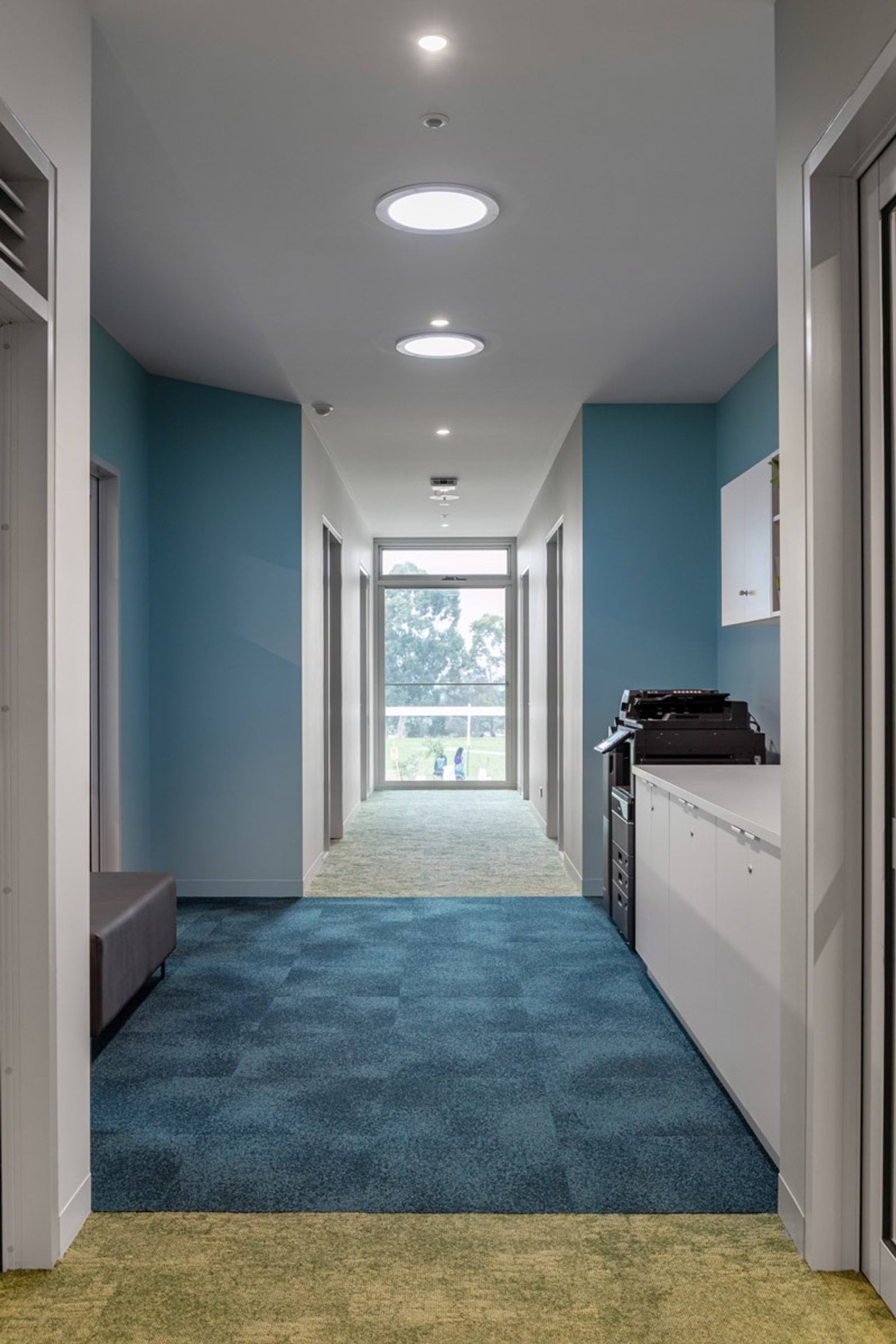 Modern school corridor with blue and green carpet, skylights, and a printer station along one wall.