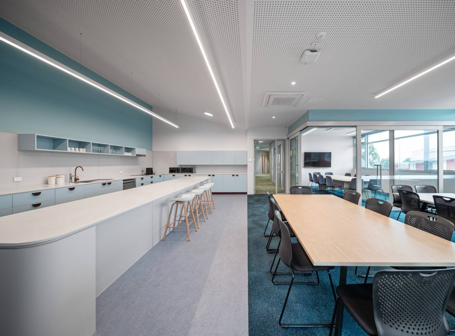 Spacious staffroom with long kitchen bench, stools, and adjoining meeting area with tables and chairs.