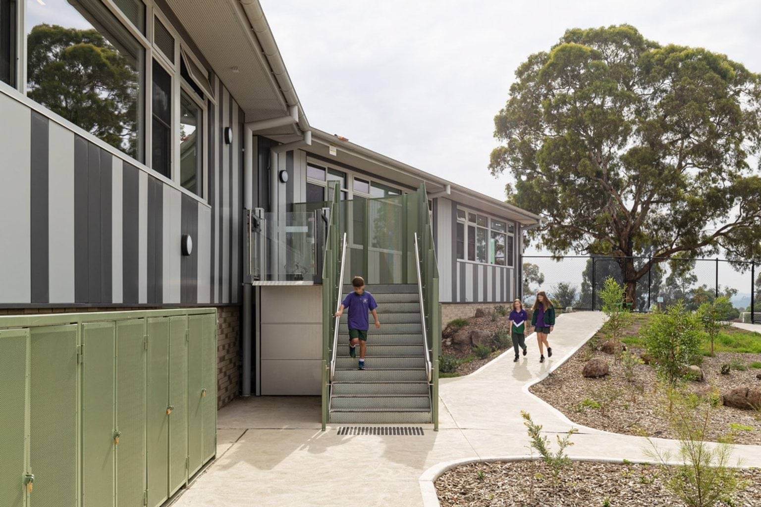 Students walking near a school building with striped cladding, outdoor stairs, and landscaped garden beds.