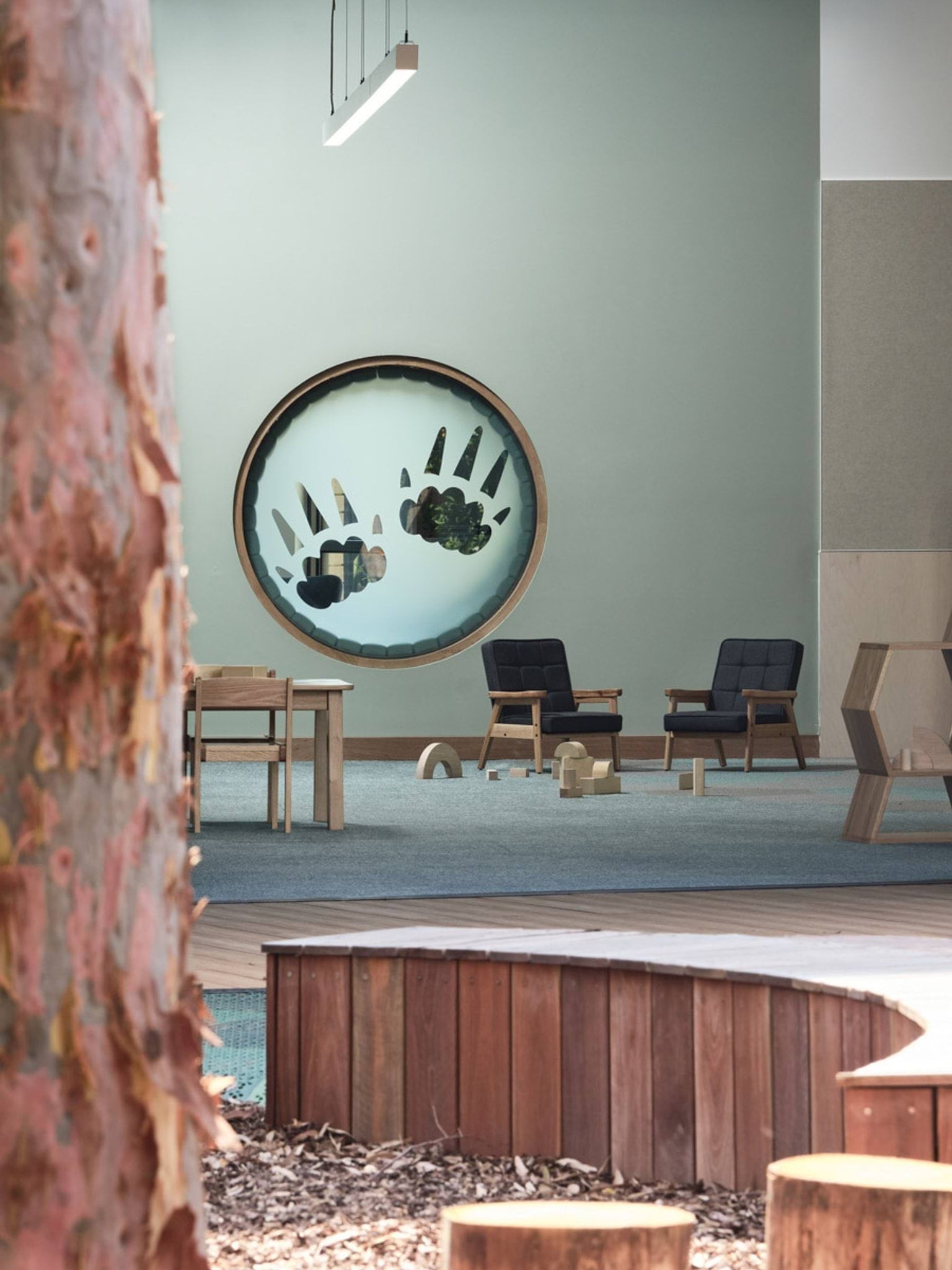 A view from the outdoor play area into the kindergarten classroom through large glass doors. Timber tables and chairs are arranged inside, and a tricycle sits on the green play mat outside.