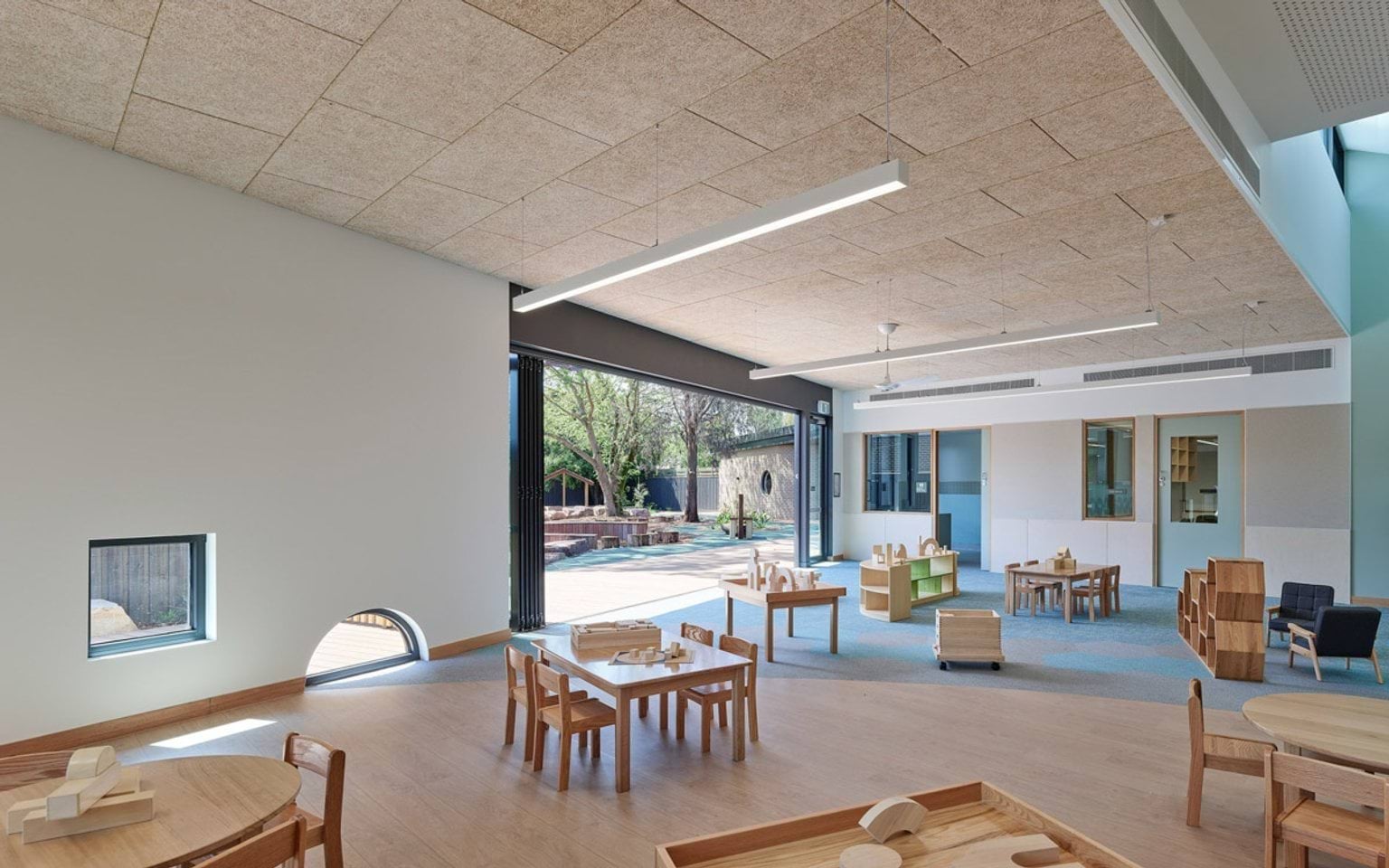 A curved section of the kindergarten exterior with cream brick walls and a round window. Timber decking and green play mats lead to a black fence.