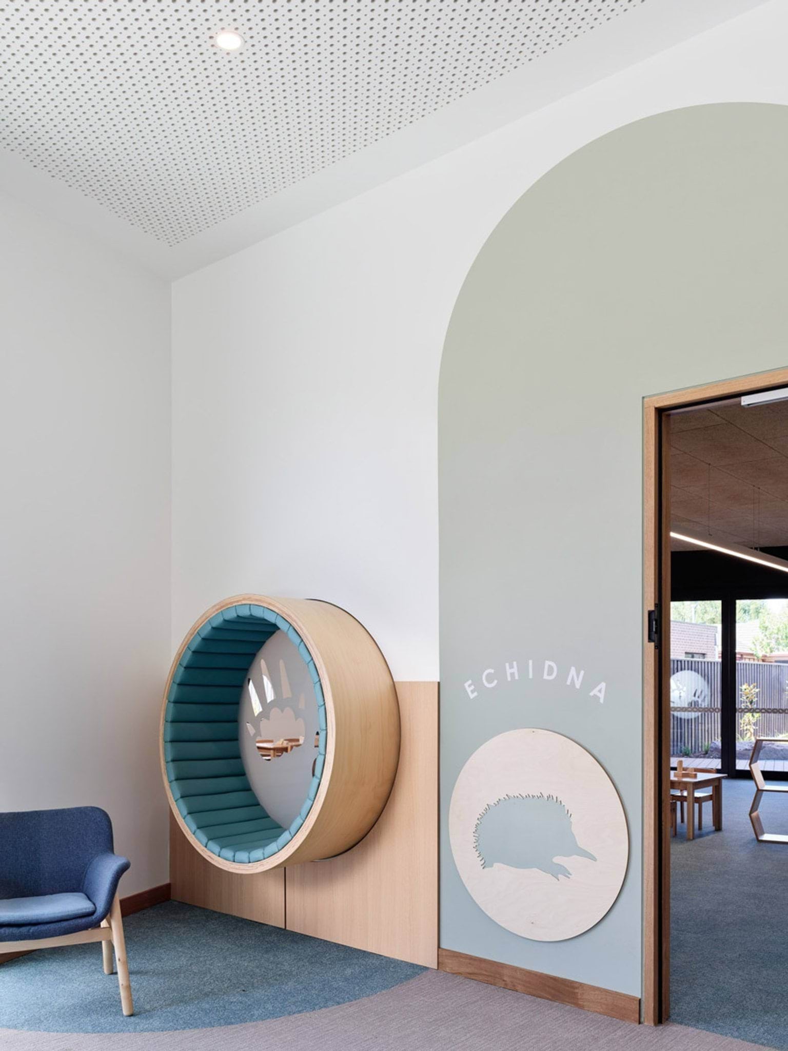 A circular timber seating nook built into a wall, with soft blue cushions and an animal footprint design. A blue chair sits nearby and a sign reads “Echidna”.