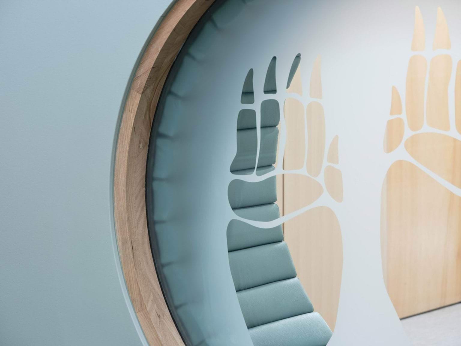 A circular timber seating nook built into a pale green wall, with soft blue cushions. The cut-out design features stylised animal footprints.