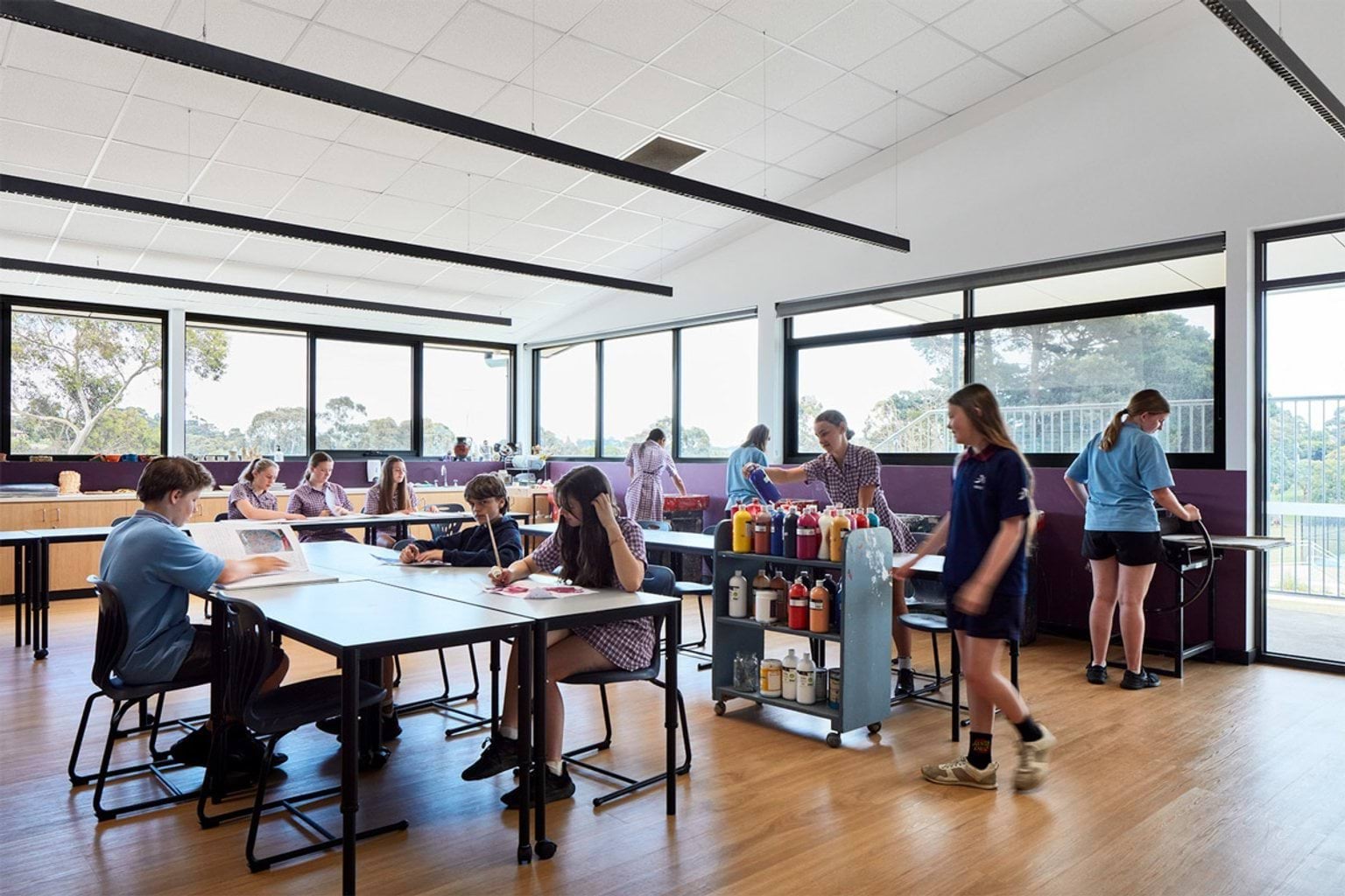 Art classroom with large windows and natural light, students seated at tables working on projects with paint supplies on a trolley