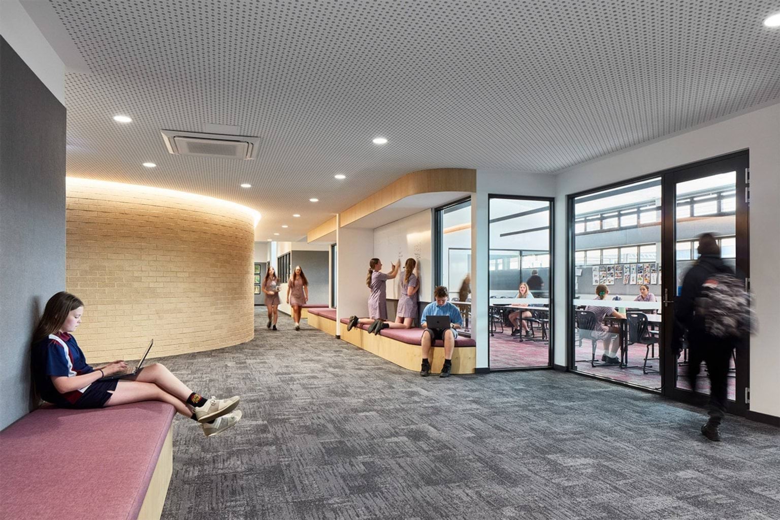 School breakout space with curved brick feature wall, cushioned bench seating and students sitting and talking near glass partitions