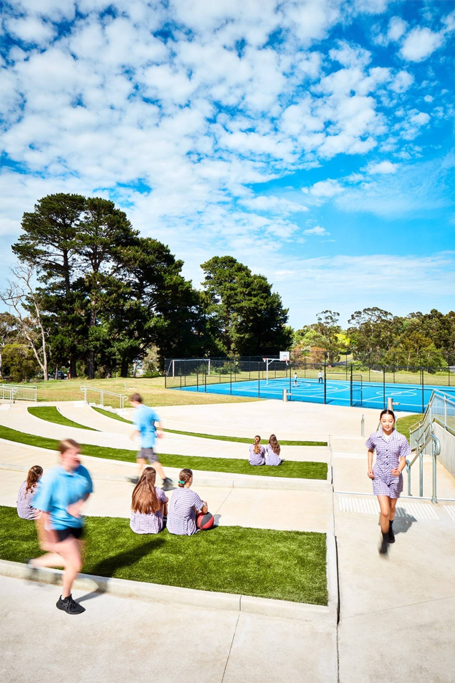Outdoor tiered seating area with concrete steps and grass, students sitting and walking near sports courts