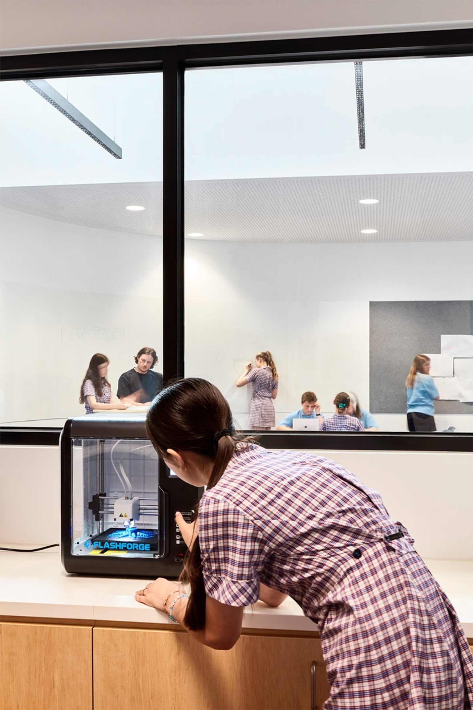 Student using a 3D printer in a classroom, with group of students working together in adjoining room visible through glass wall