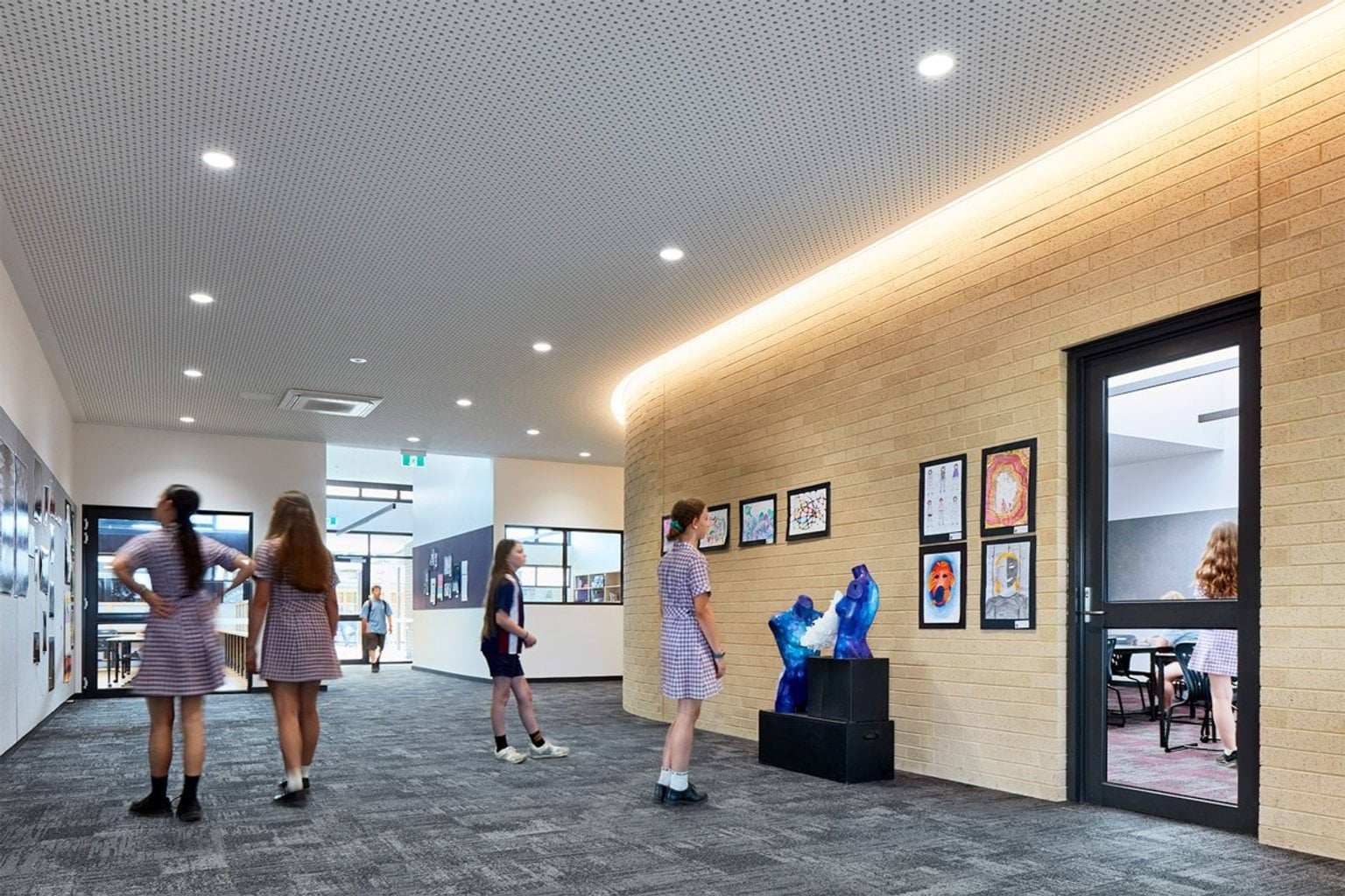 School corridor with curved brick wall, display of student artwork and sculptures, and students walking through space