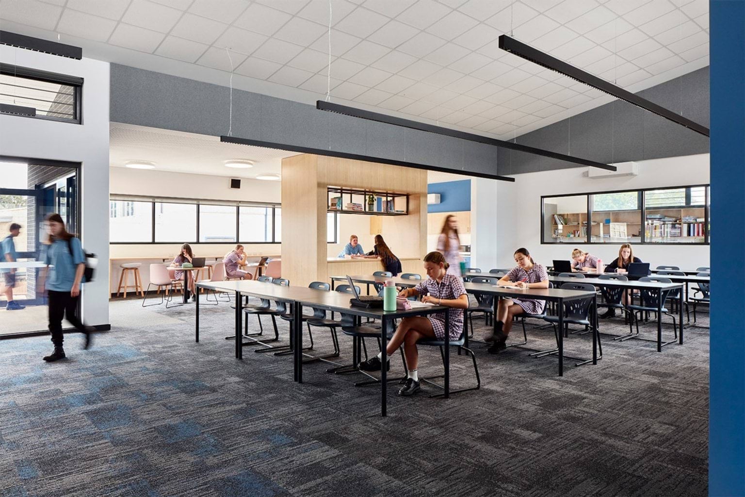 Classroom with rows of desks and chairs, central timber feature wall and students working on laptops and writing