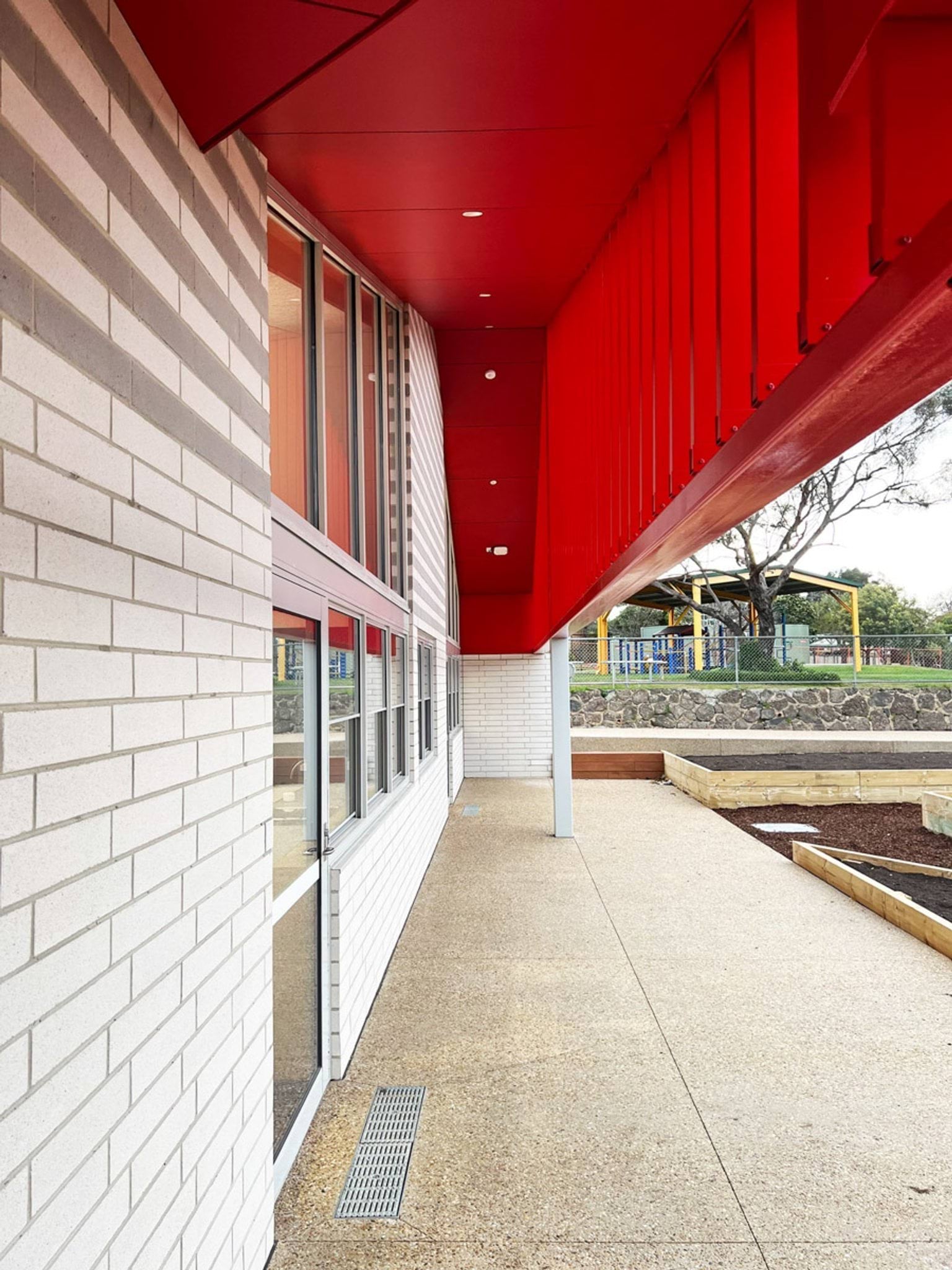 A covered outdoor area with a bold red steel canopy and white brick walls. Raised garden beds and a playground are visible in the background.