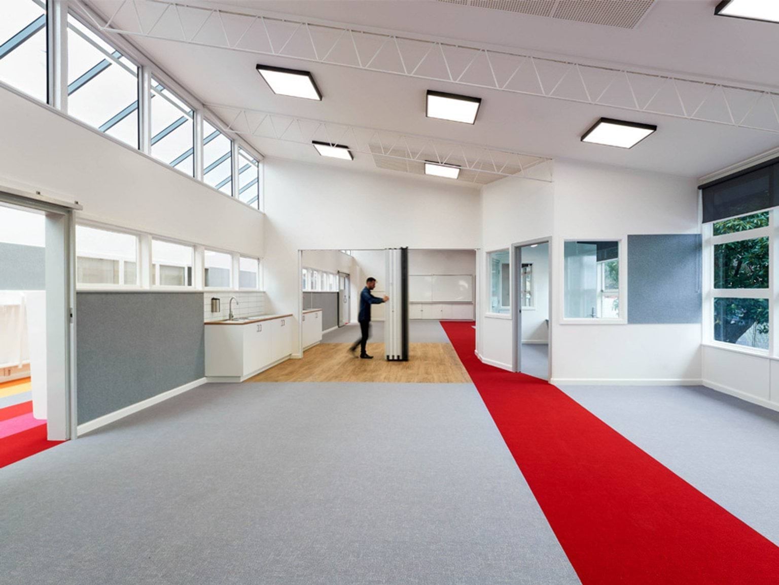 A large open learning area with grey carpet, a red walkway, and high ceilings with square lights. A person is adjusting a folding partition near a kitchenette.
