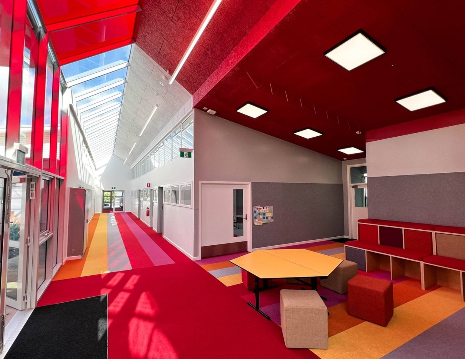 A long hallway with a glass skylight roof and colorful striped carpet in red, orange, and yellow. The walls are white with grey panels, and there are doors to classrooms along both sides.