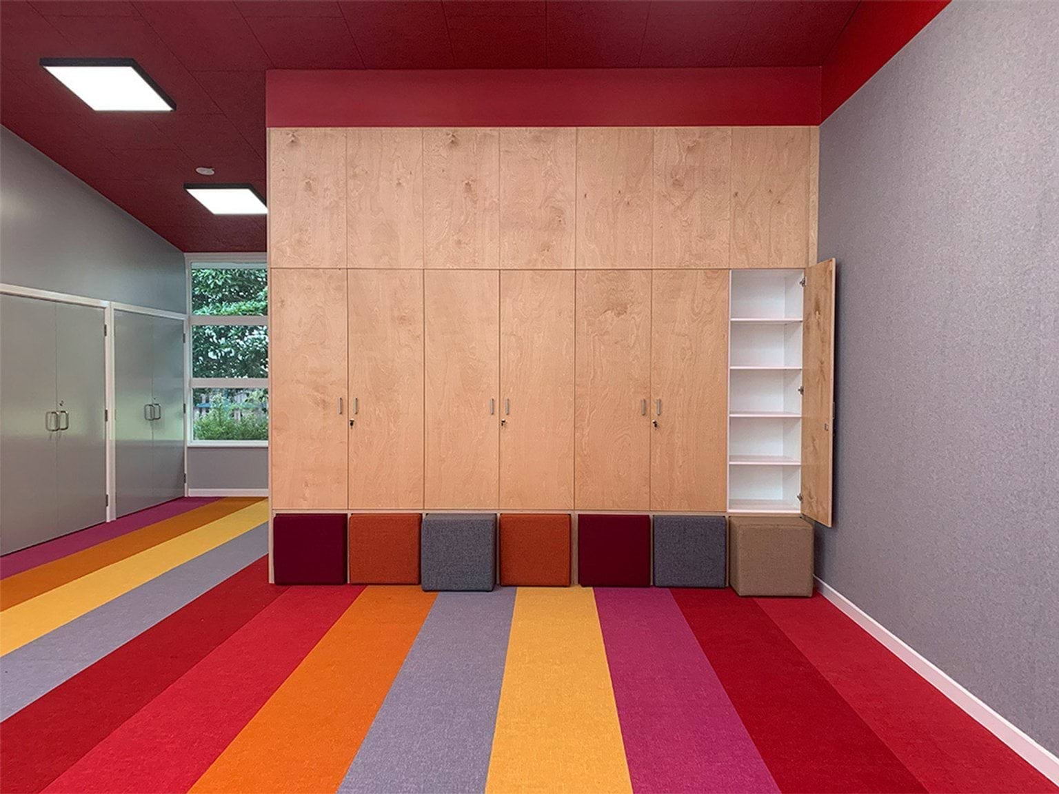 An interior space with a timber storage wall and colourful cube seating along the base. The floor has bright striped carpet in red, orange, and purple.