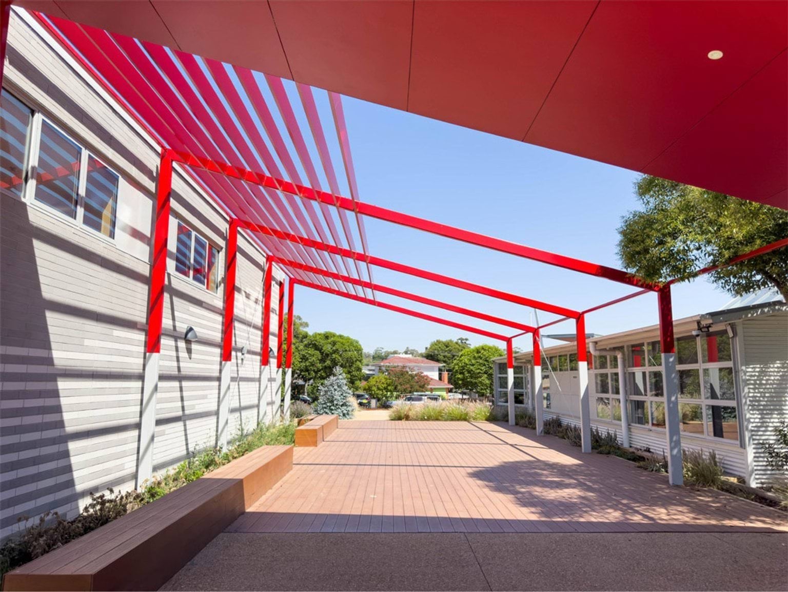 An outdoor walkway with a striking red steel canopy and timber seating. Landscaping with shrubs and garden beds surrounds the area.
