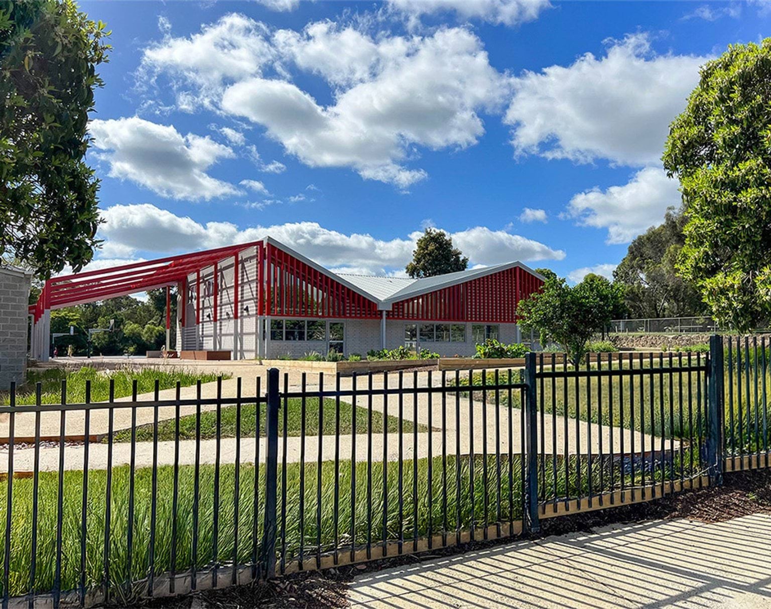 The front view of a modern school building with angled roofs and red steel frames, seen behind a black metal fence. Landscaping and pathways are in the foreground.