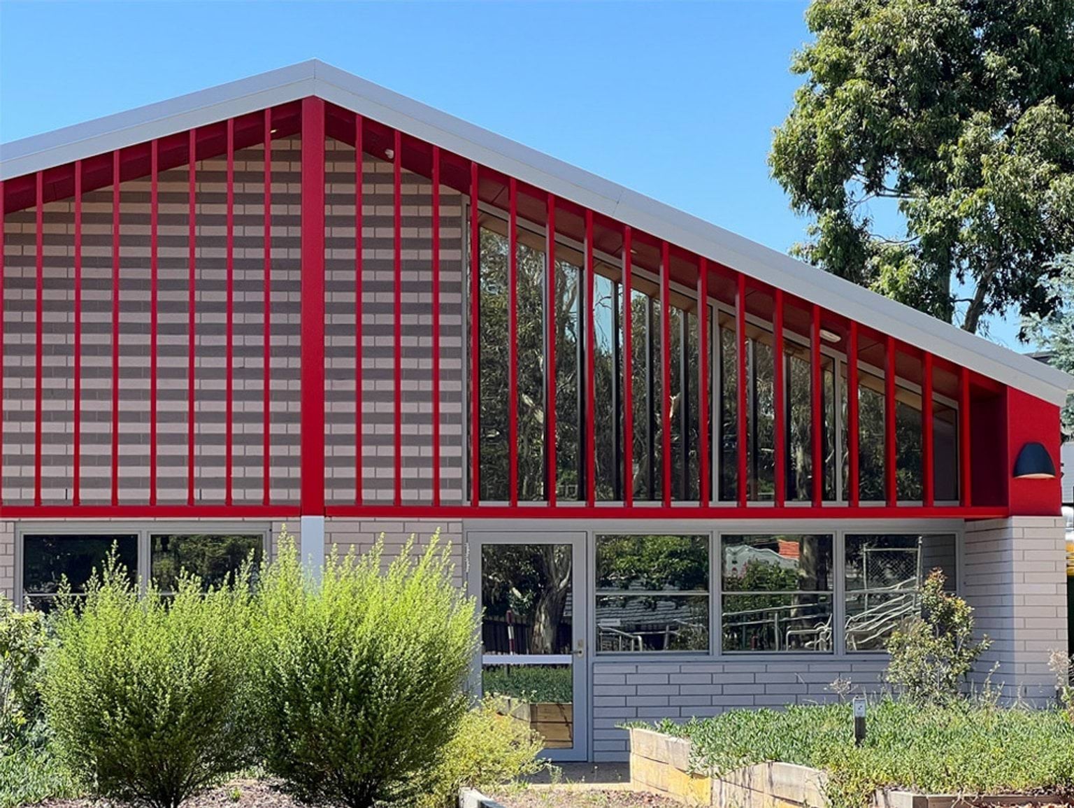 The exterior of a school building with a sloped roof and bold red vertical frames over large windows. Green landscaping is in front of the building.