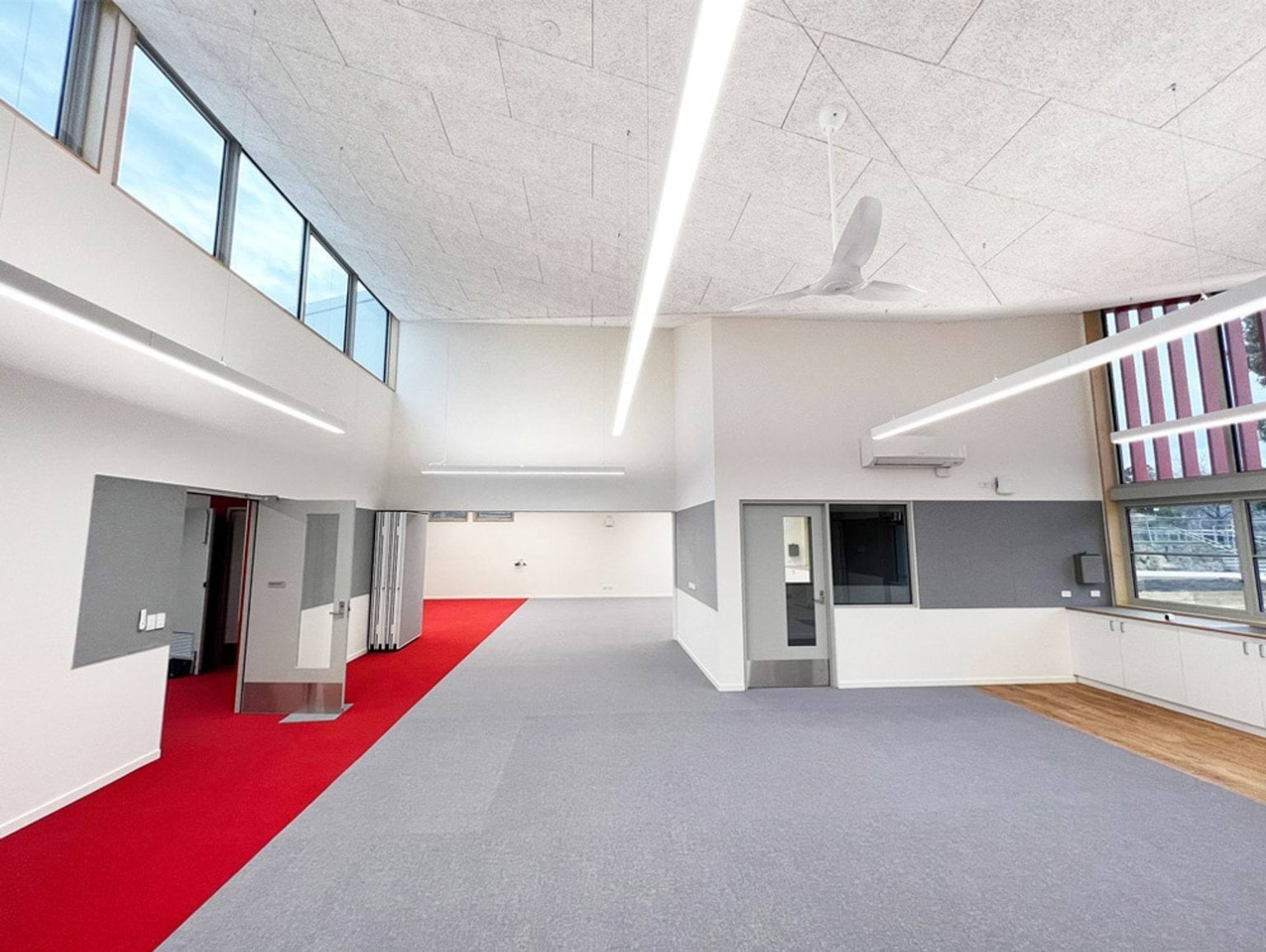 A large open classroom with grey carpet, a red walkway, and high ceilings with exposed trusses and square lights. A kitchenette is in one corner.