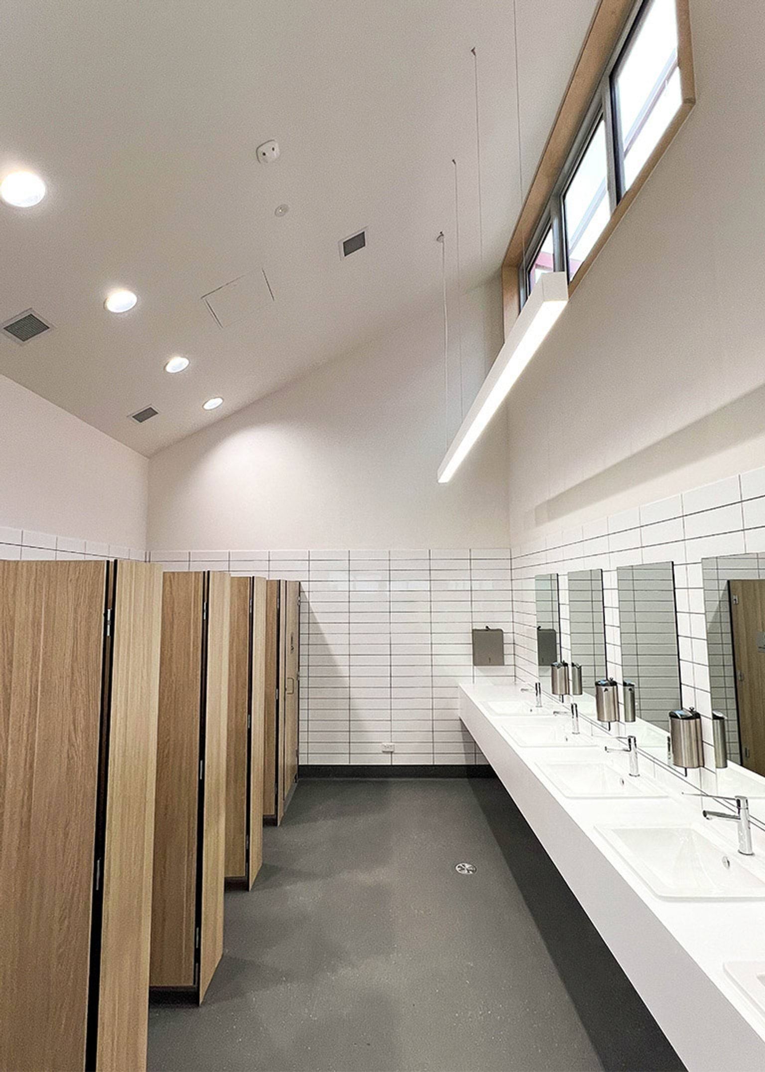 A bathroom with timber cubicle doors, white tiled walls, and a long vanity with multiple sinks and mirrors under a high ceiling.