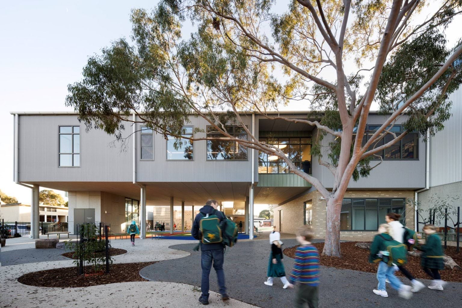 A landscaped school courtyard with curved paths, garden beds, and a large tree. Students and an adult walk towards the building.