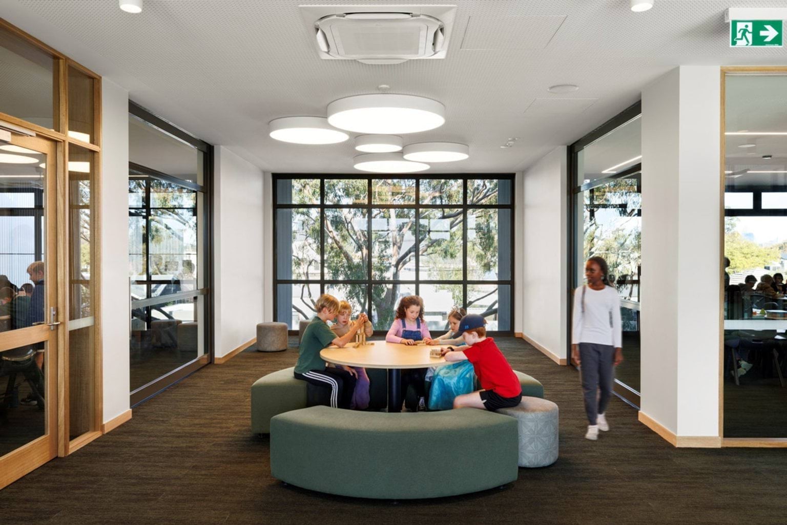 Students sitting around a circular table in a light-filled learning space with large windows and round ceiling lights.