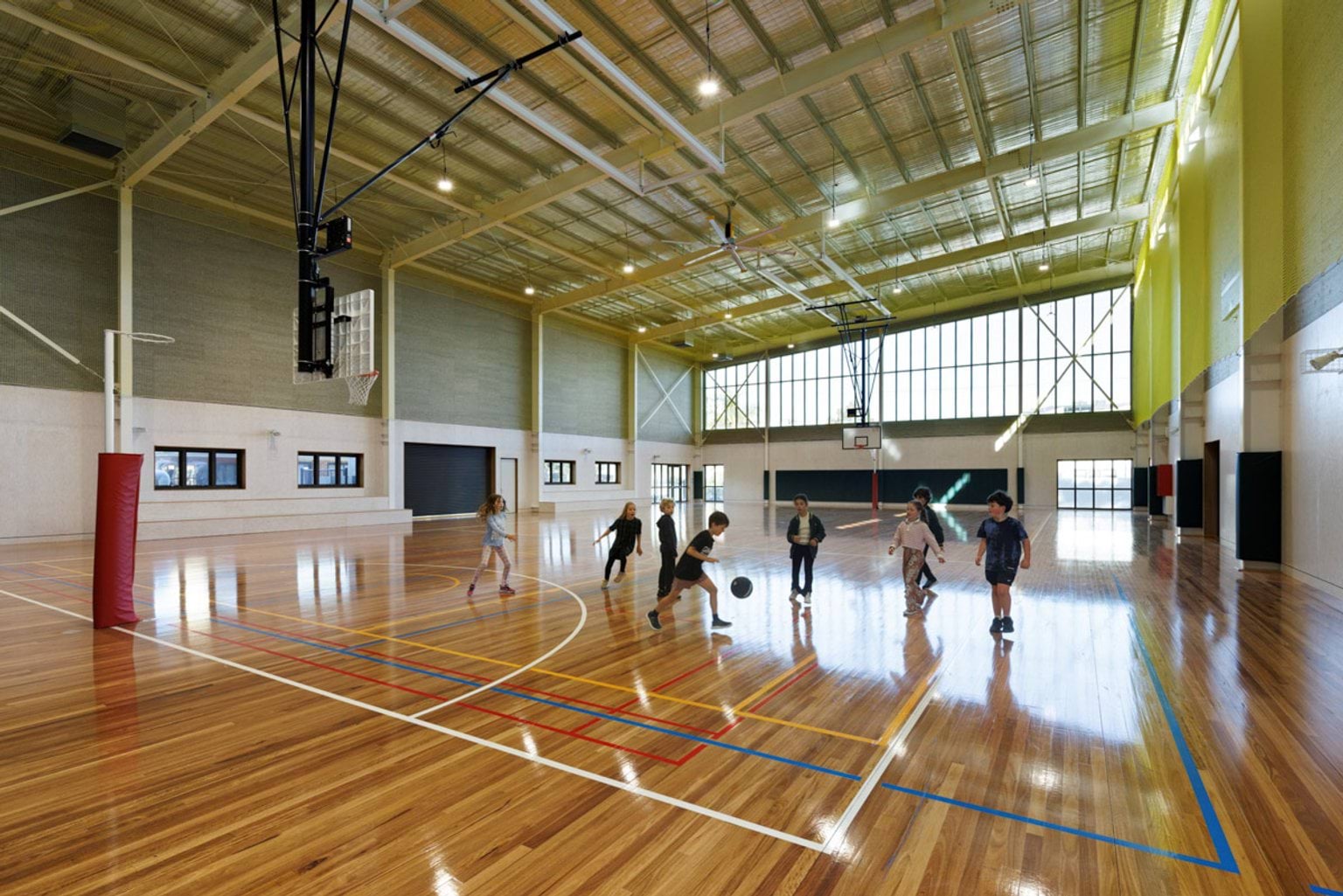 A spacious indoor sports hall with polished timber flooring, high ceilings, and basketball hoops. Students are playing ball games.