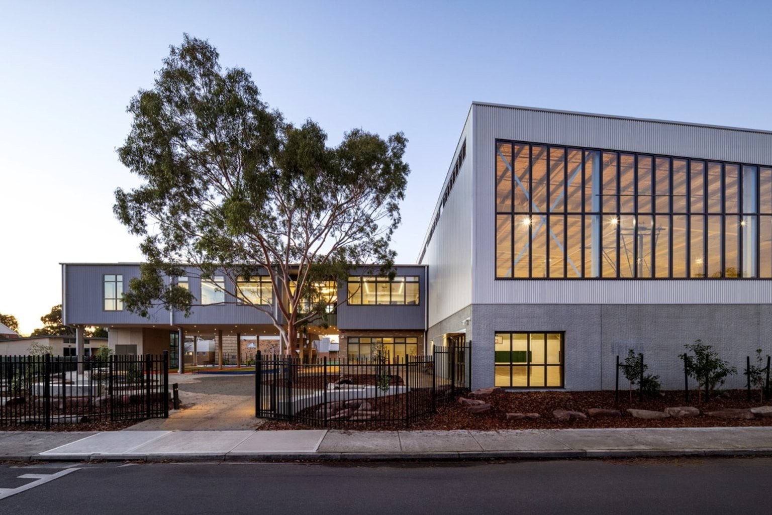 The exterior of a school building with tall glass windows and landscaped garden beds, illuminated in the evening.