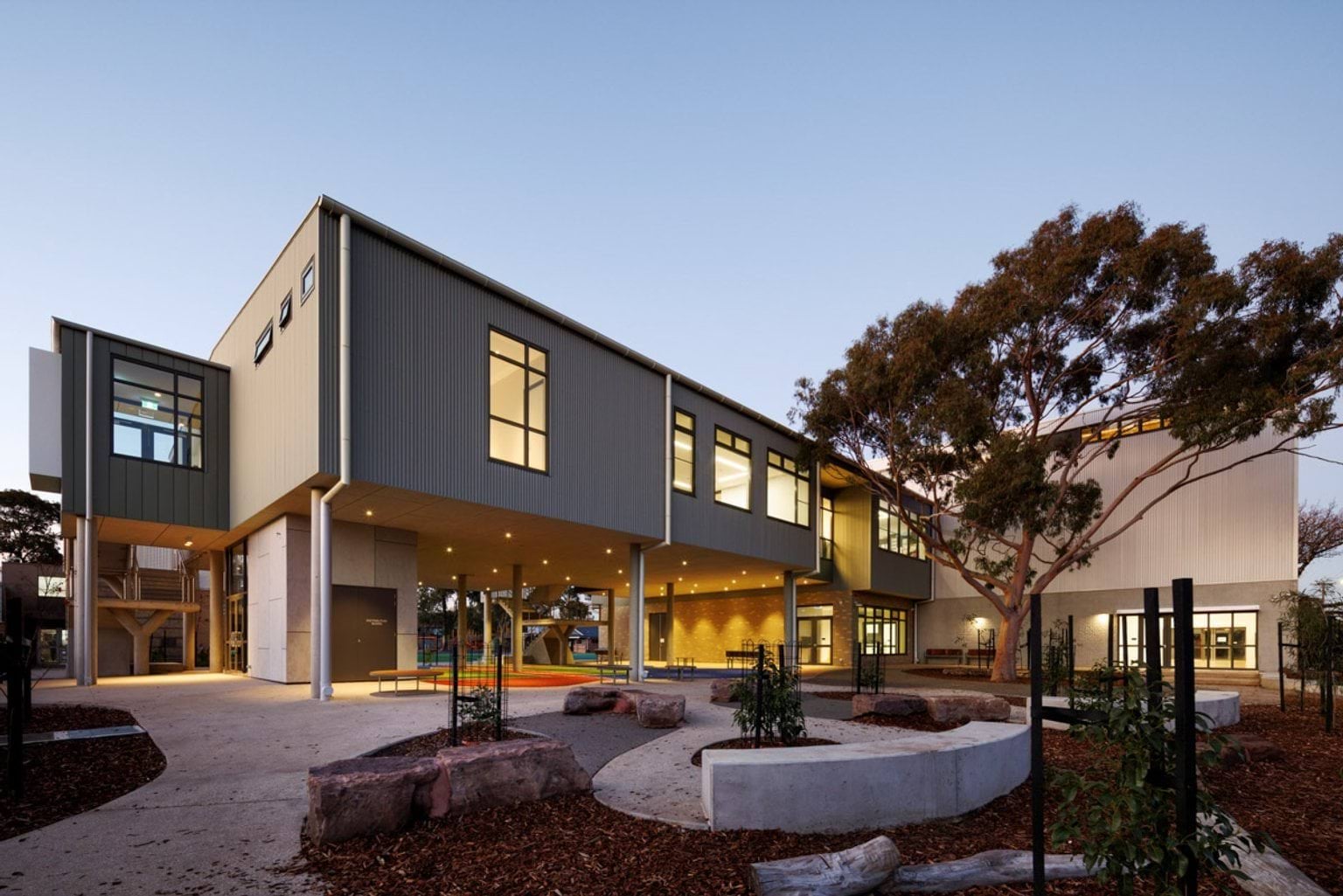 A two-storey school building with large windows and an open undercroft, illuminated at dusk. Landscaping includes trees and garden beds.