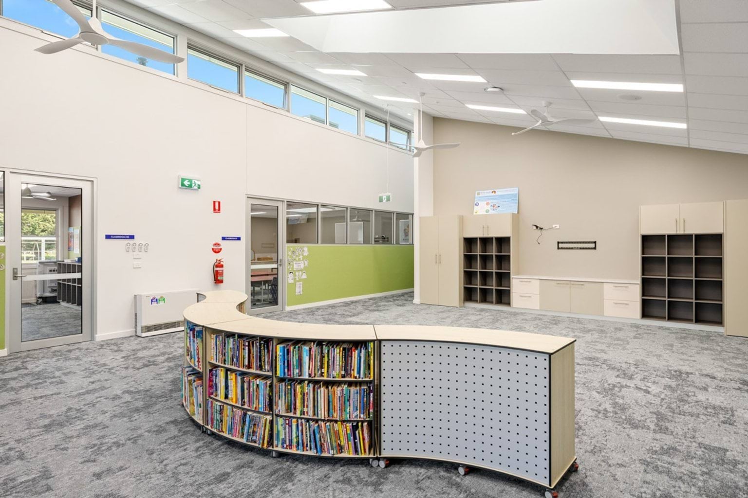 A bright library space with curved shelving filled with books, grey carpet, and high windows. Built-in storage cabinets line the back wall.