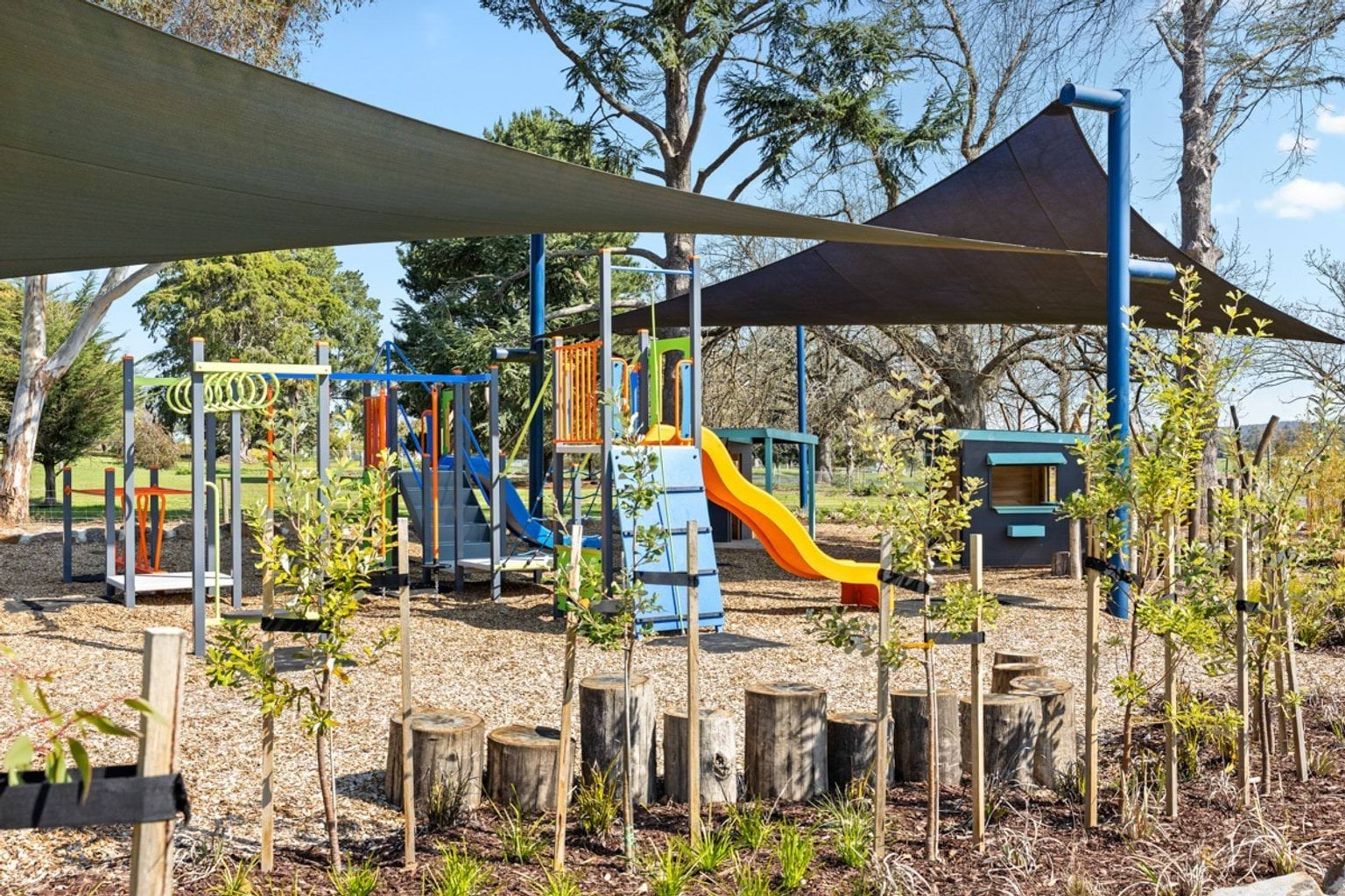 An outdoor playground with colourful slides, climbing frames, and shade sails. The ground is covered in wood chips.