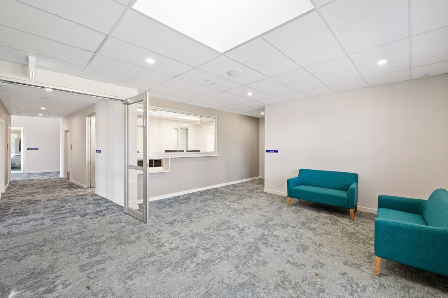 A reception area with two teal lounge chairs, grey carpet, and a glass door leading to an office counter with a large window.